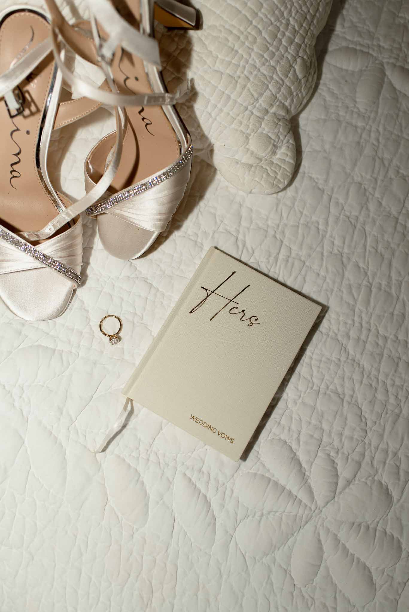 Flat lay of ivory crystal-strap heels, wedding vows booklet, and gold diamond ring on white quilt