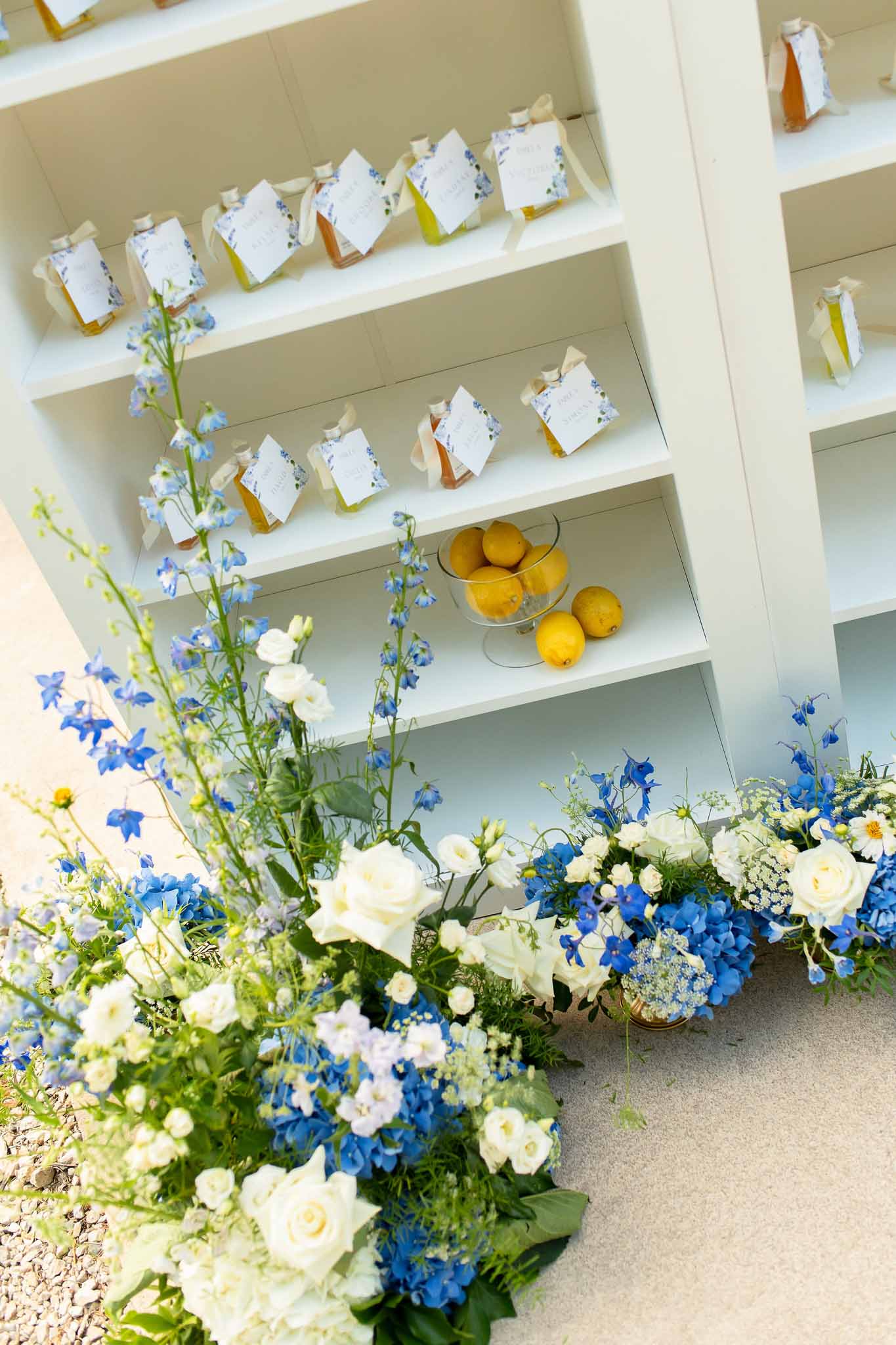 Honey bottle wedding favors on white shelf with blue hydrangea and delphinium arrangements below