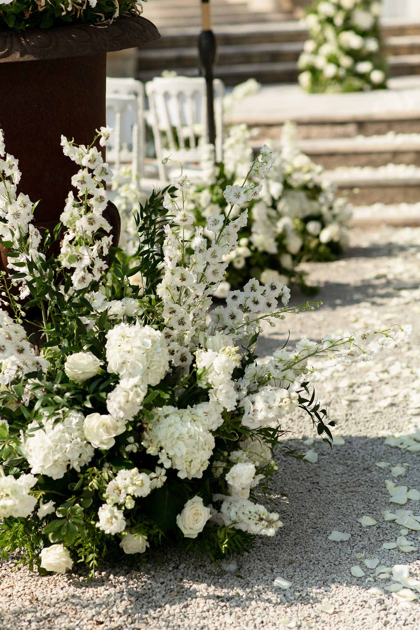 White hydrangea and rose floral arrangements lining a gravel ceremony aisle with scattered petals and Chiavari chairs