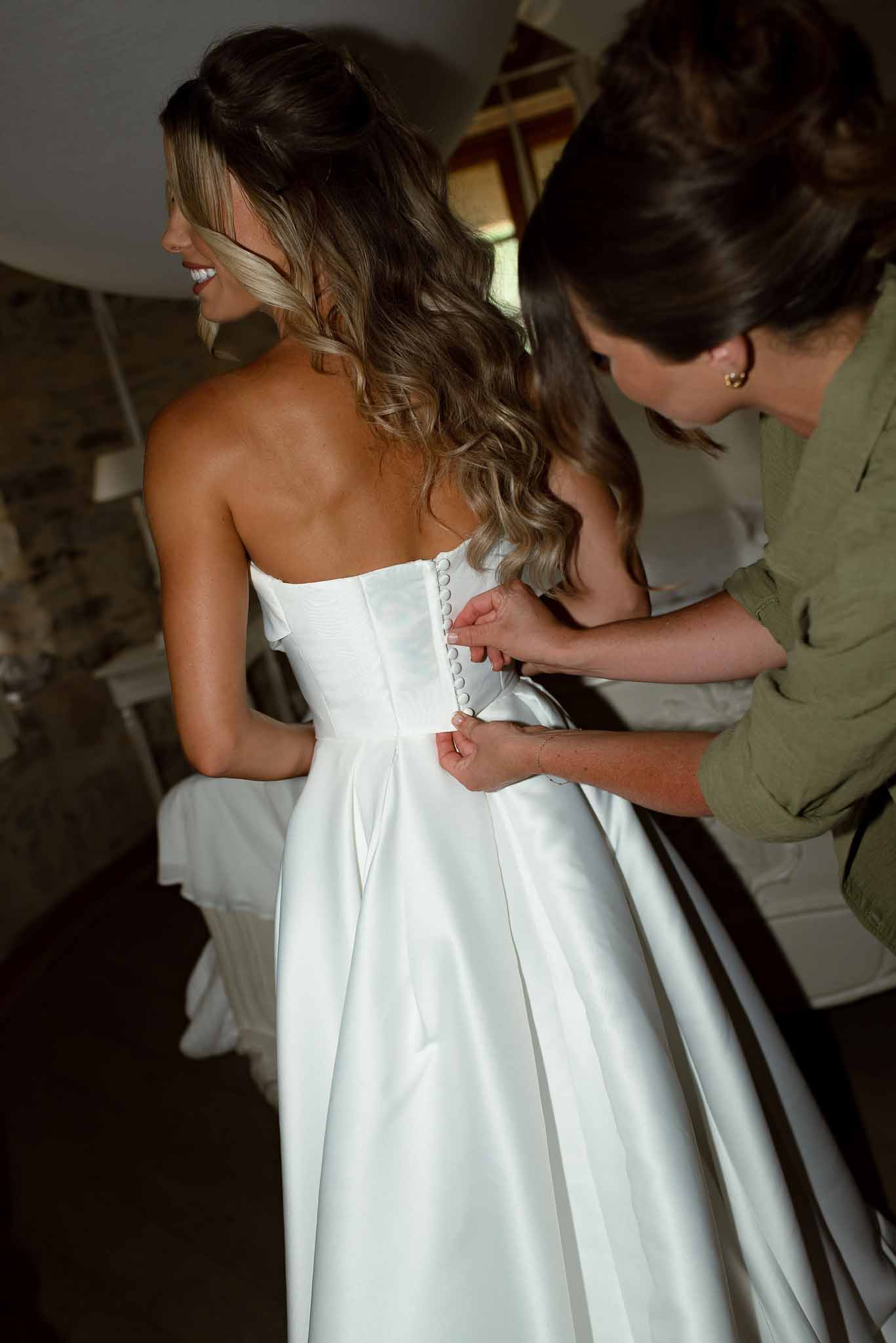 Woman fastening back buttons of bride's strapless satin ballgown in dimly lit stone-walled chateau room