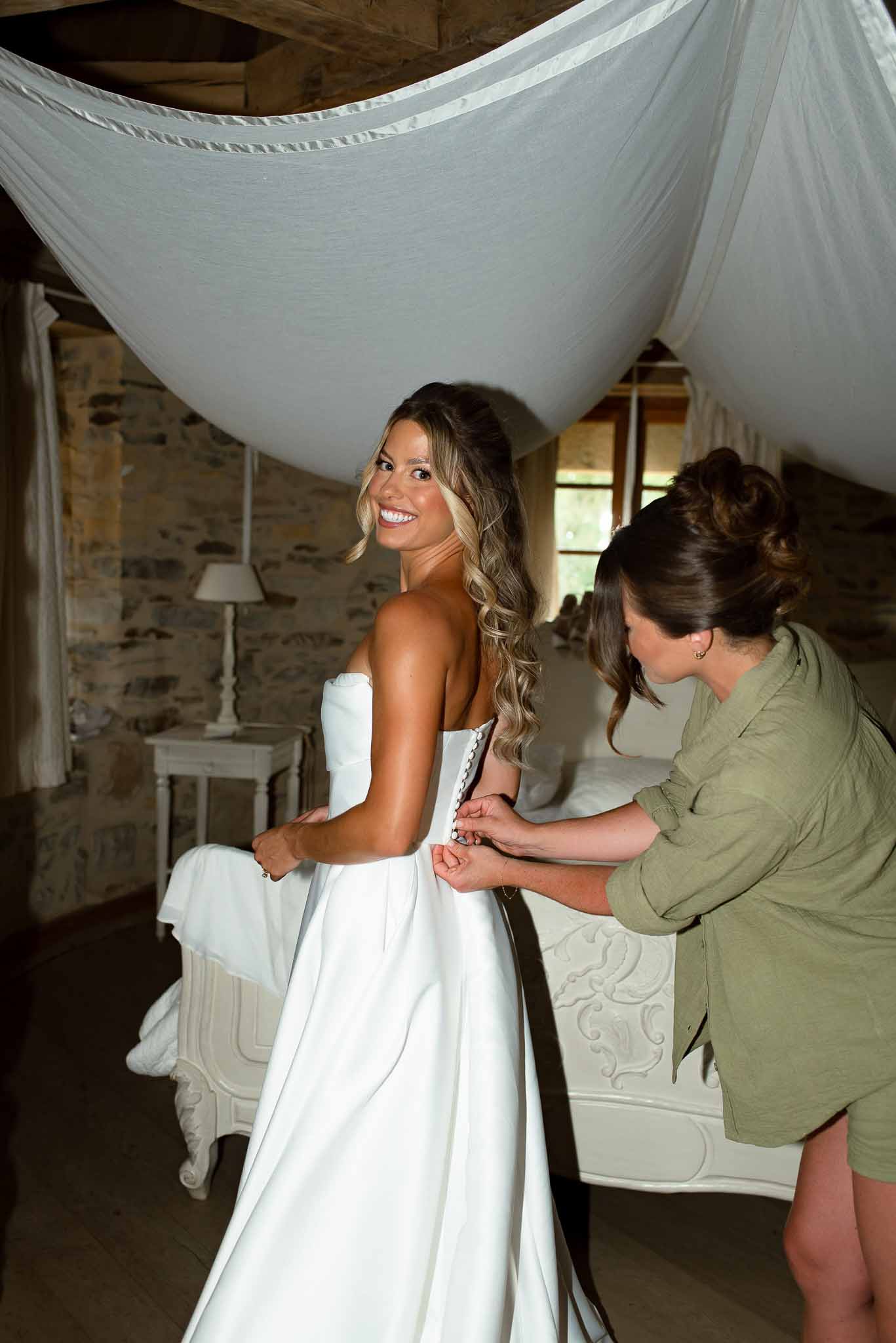 Smiling bride having her strapless gown buttoned in a rustic chateau bridal suite with exposed beams and stone walls