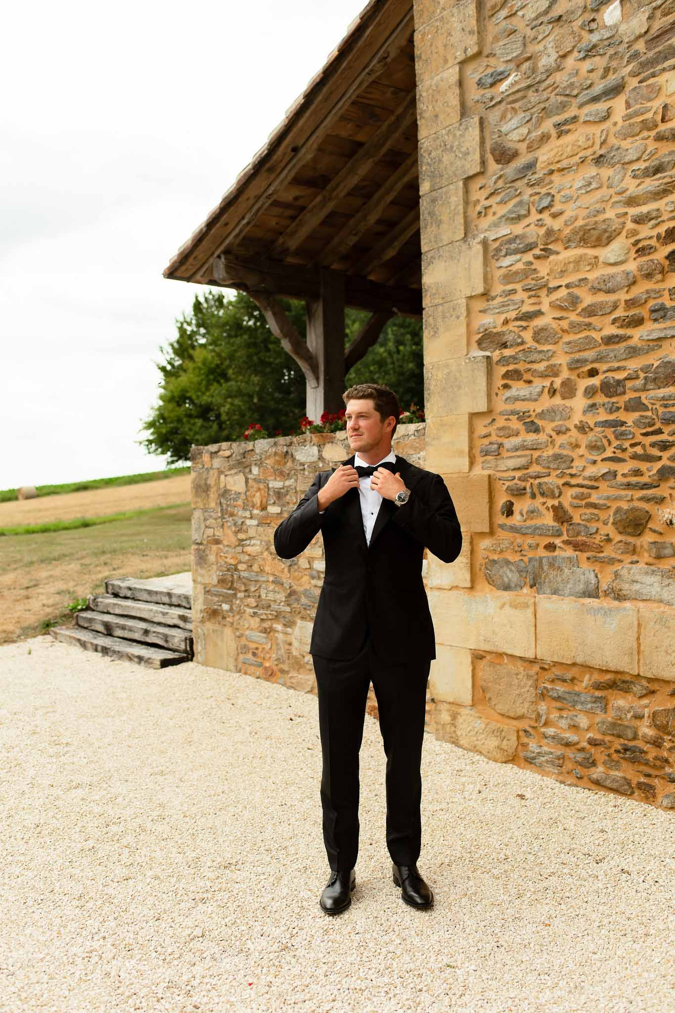 Groom adjusting black bow tie in tuxedo beside honey-toned stone building with wooden overhang