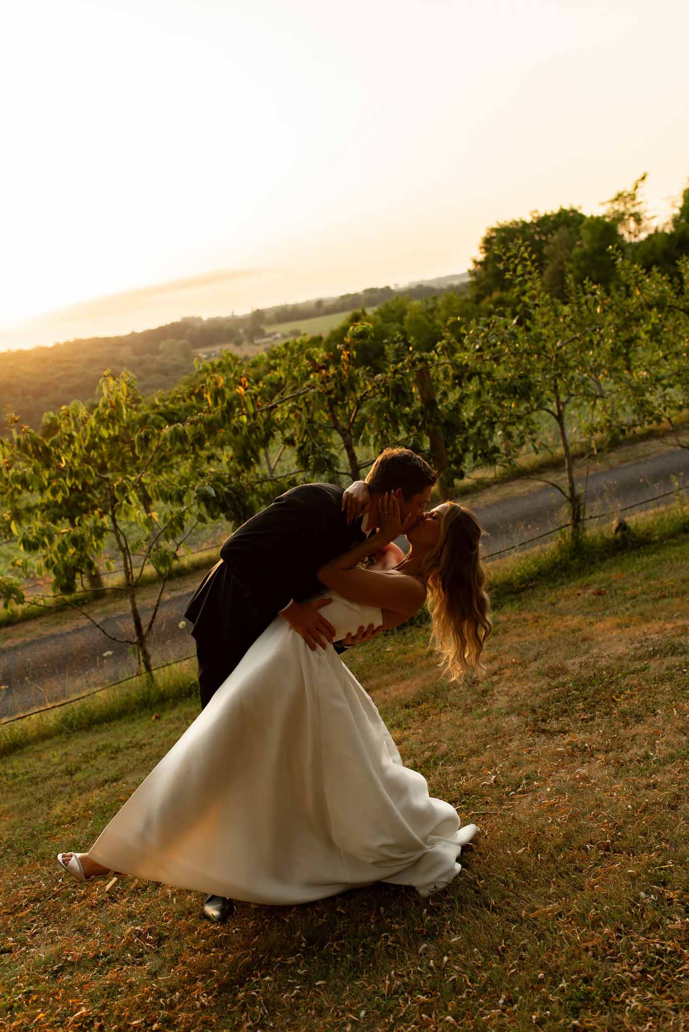 Groom dipping bride for kiss on hillside orchard at golden hour with rolling landscape