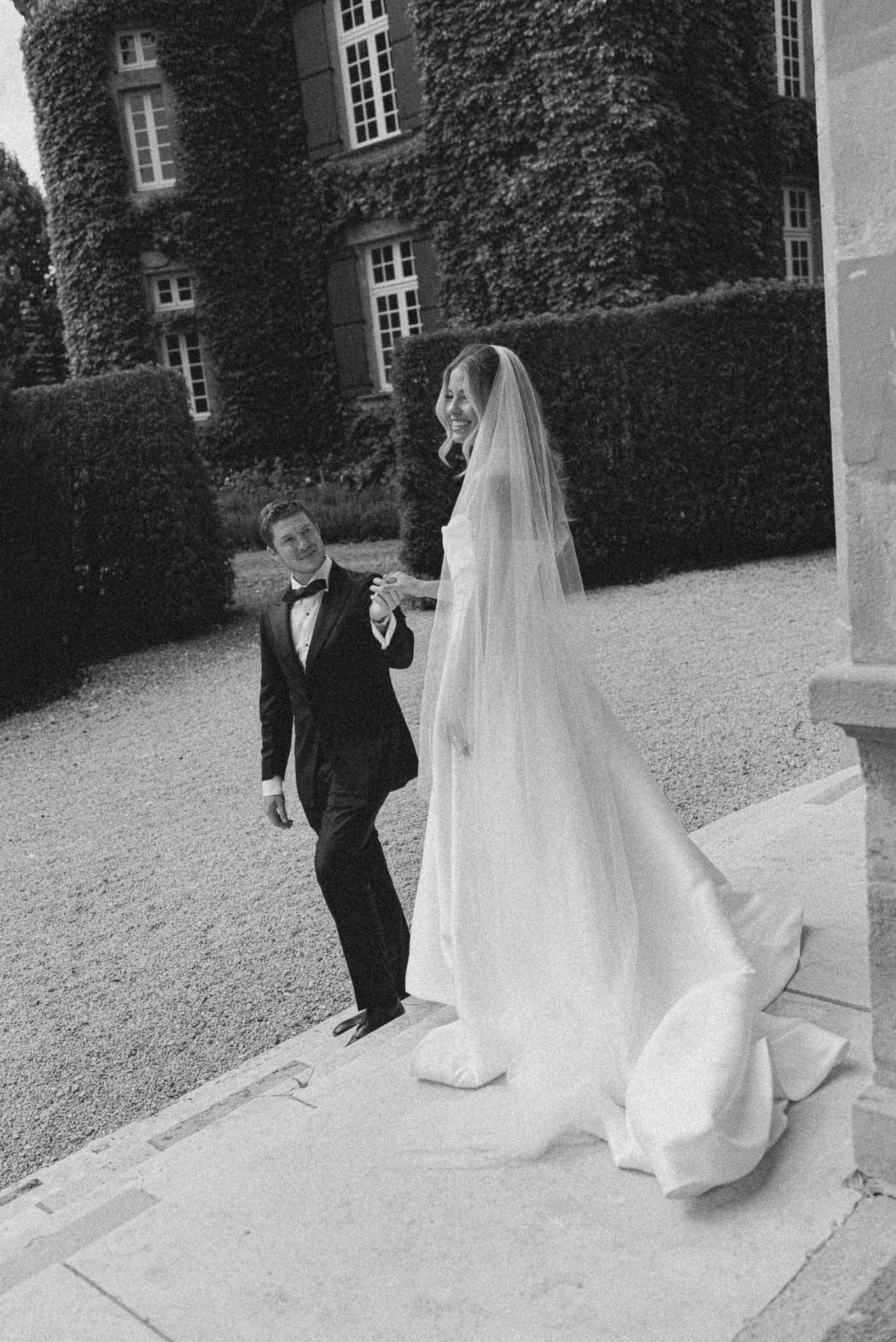 Bride with cathedral veil and dramatic train smiling at groom on ivy-covered chateau steps in B&W