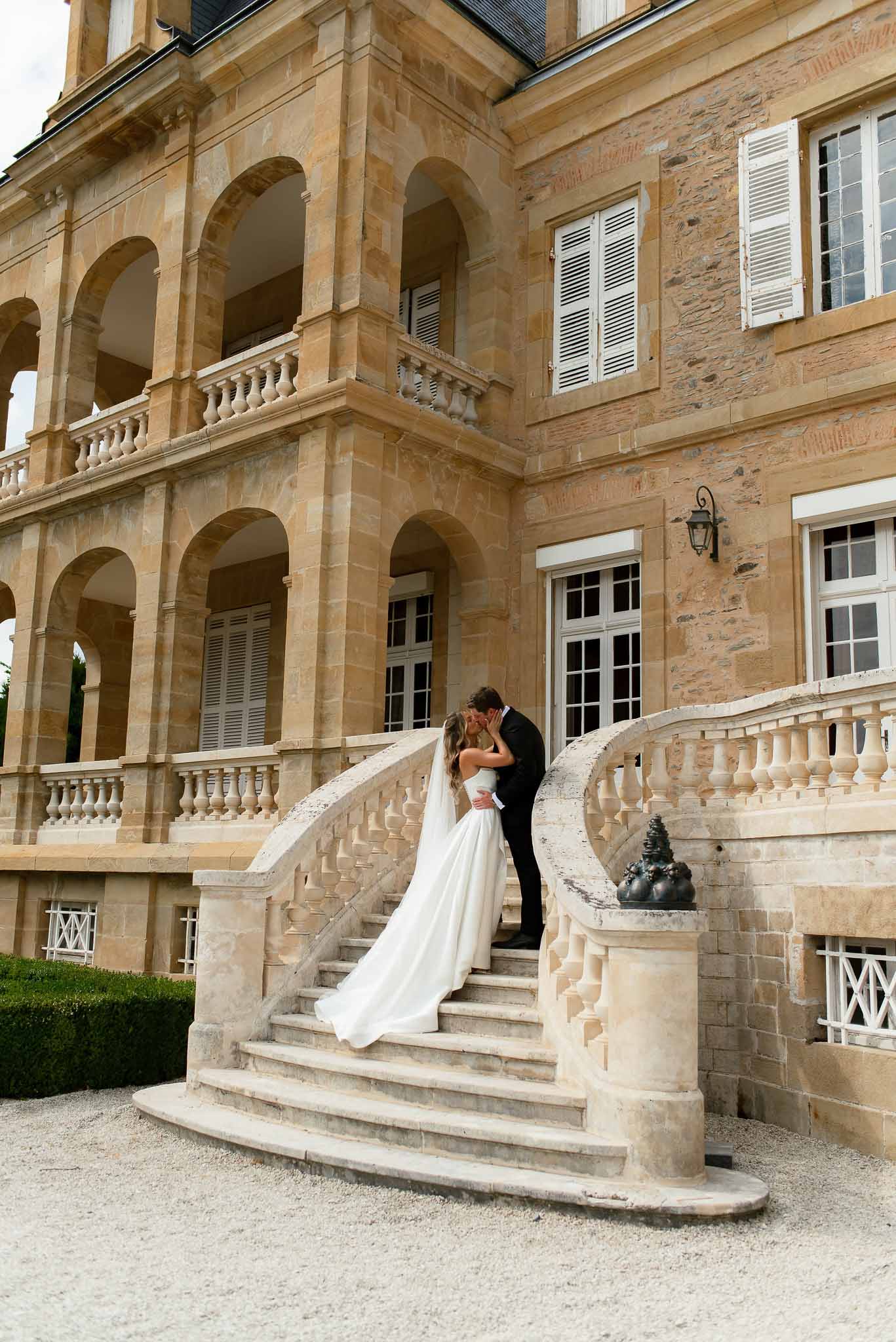 Bride and groom kissing on curved stone staircase of golden limestone chateau with colonnaded galleries