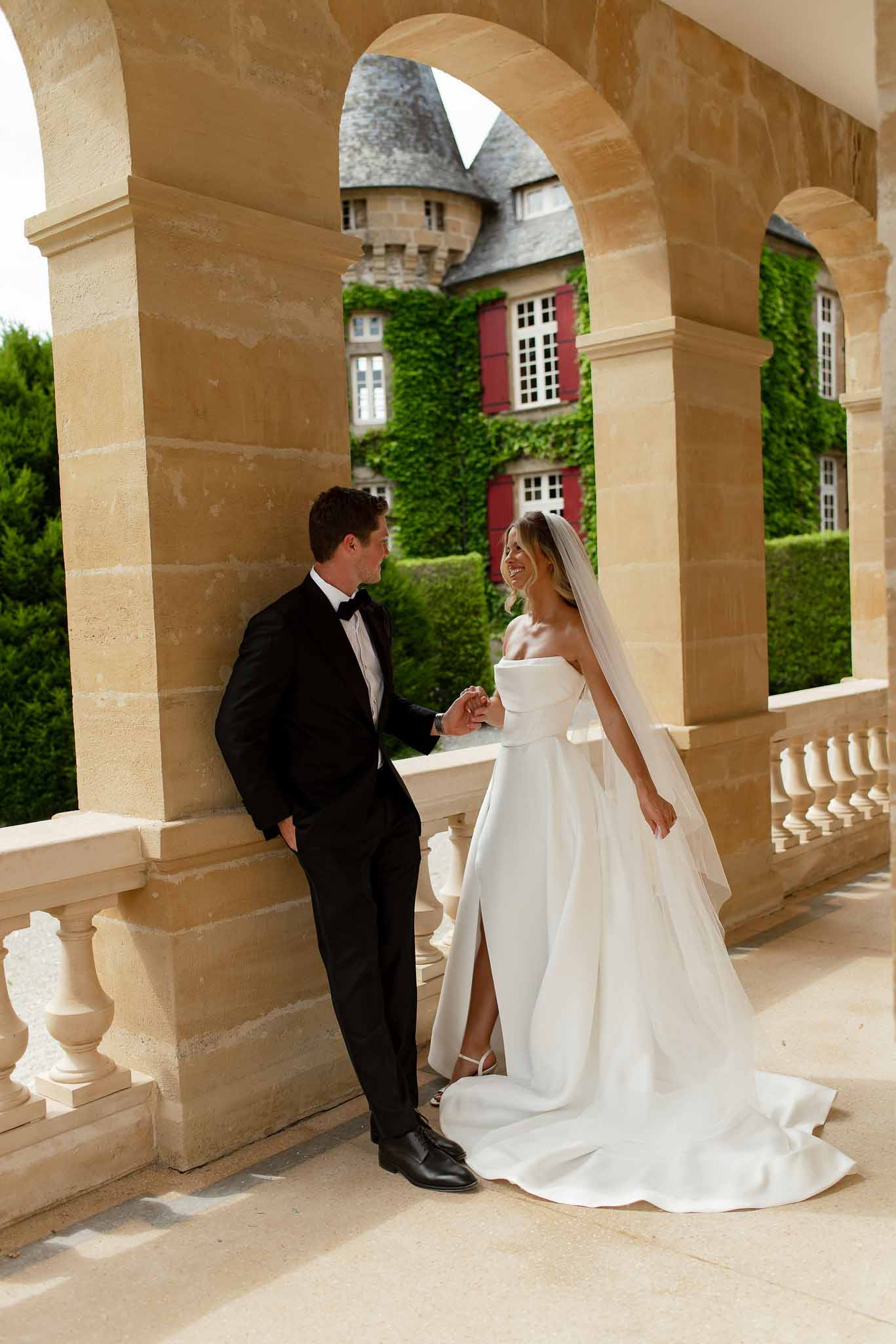 Bride in white ballgown and groom in black tuxedo laughing on chateau stone terrace with arched colonnade
