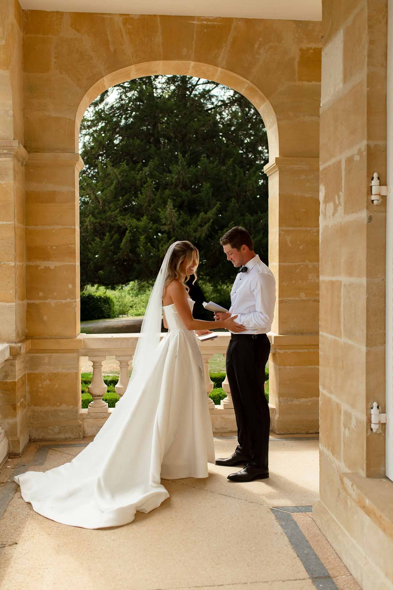 Bride and groom reading private vows on a chateau stone portico framed by a sandstone arch overlooking formal gardens