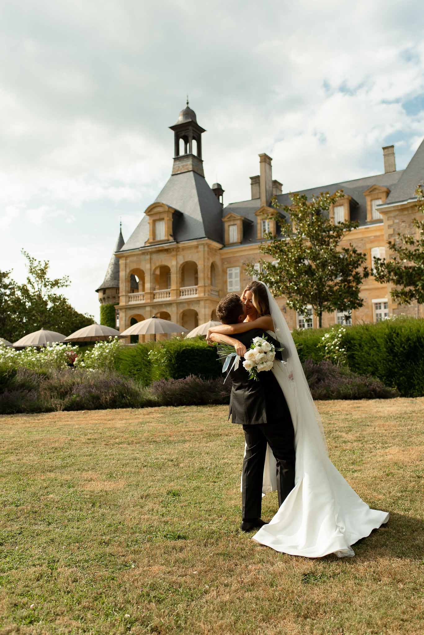 Groom lifting bride with cathedral veil and white peony bouquet on lawn before towered stone chateau