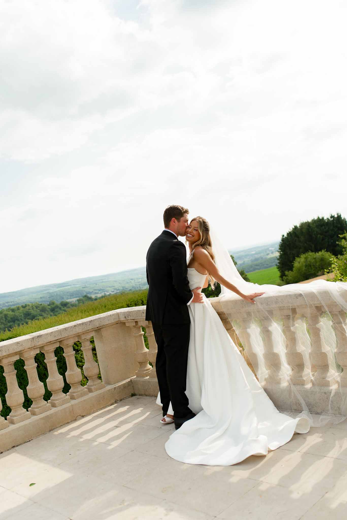 Groom kissing bride on cheek on stone terrace with rolling countryside and cathedral veil in wind