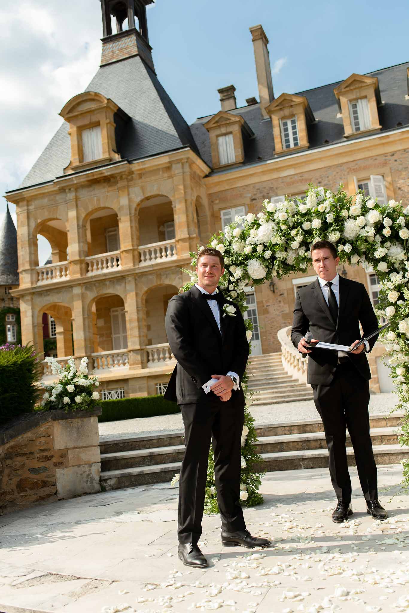 Groom and officiant beneath circular white rose arch at chateau terrace ceremony with scattered petals