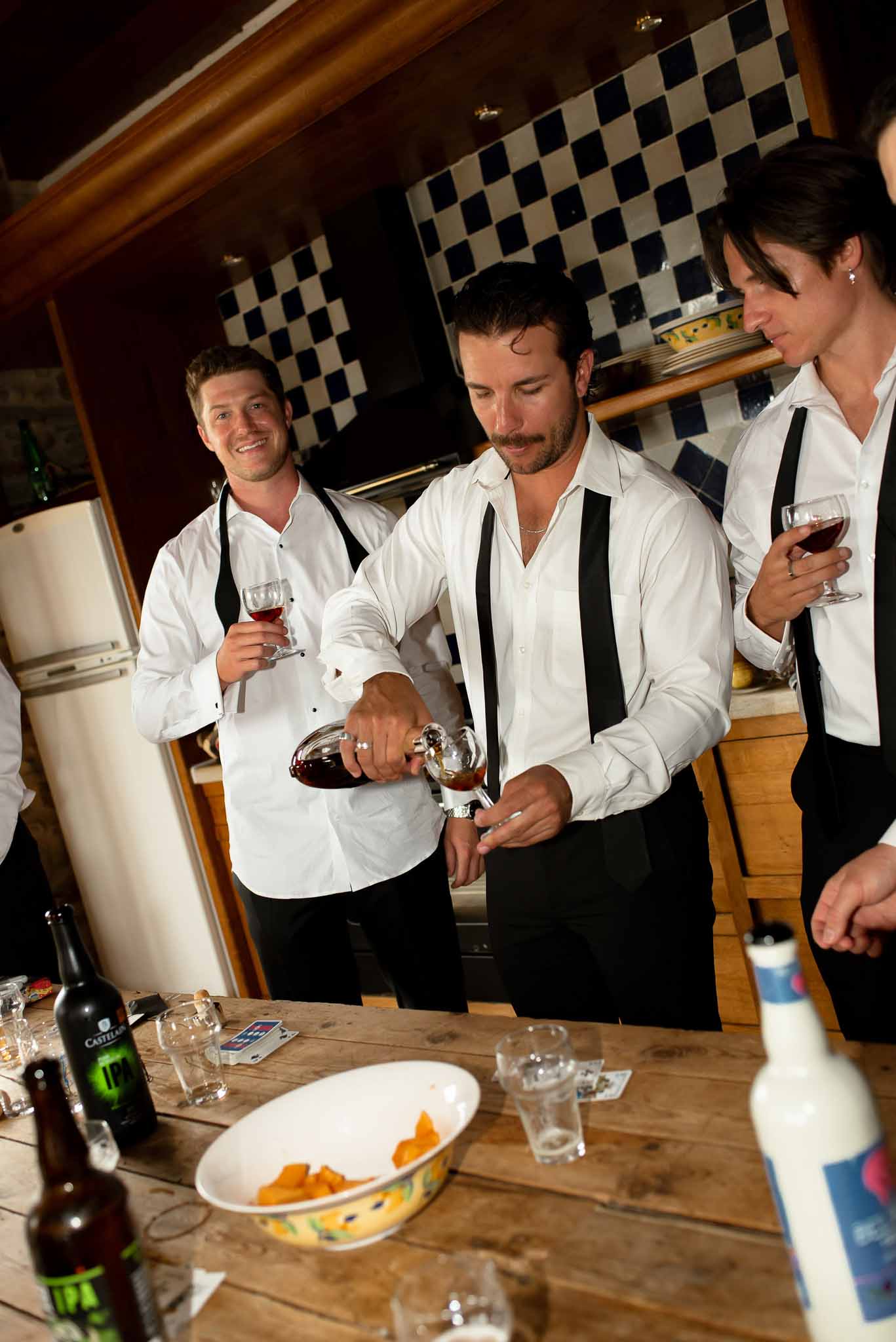 Three groomsmen in white shirts and suspenders pouring red wine in rustic farmhouse kitchen before ceremony