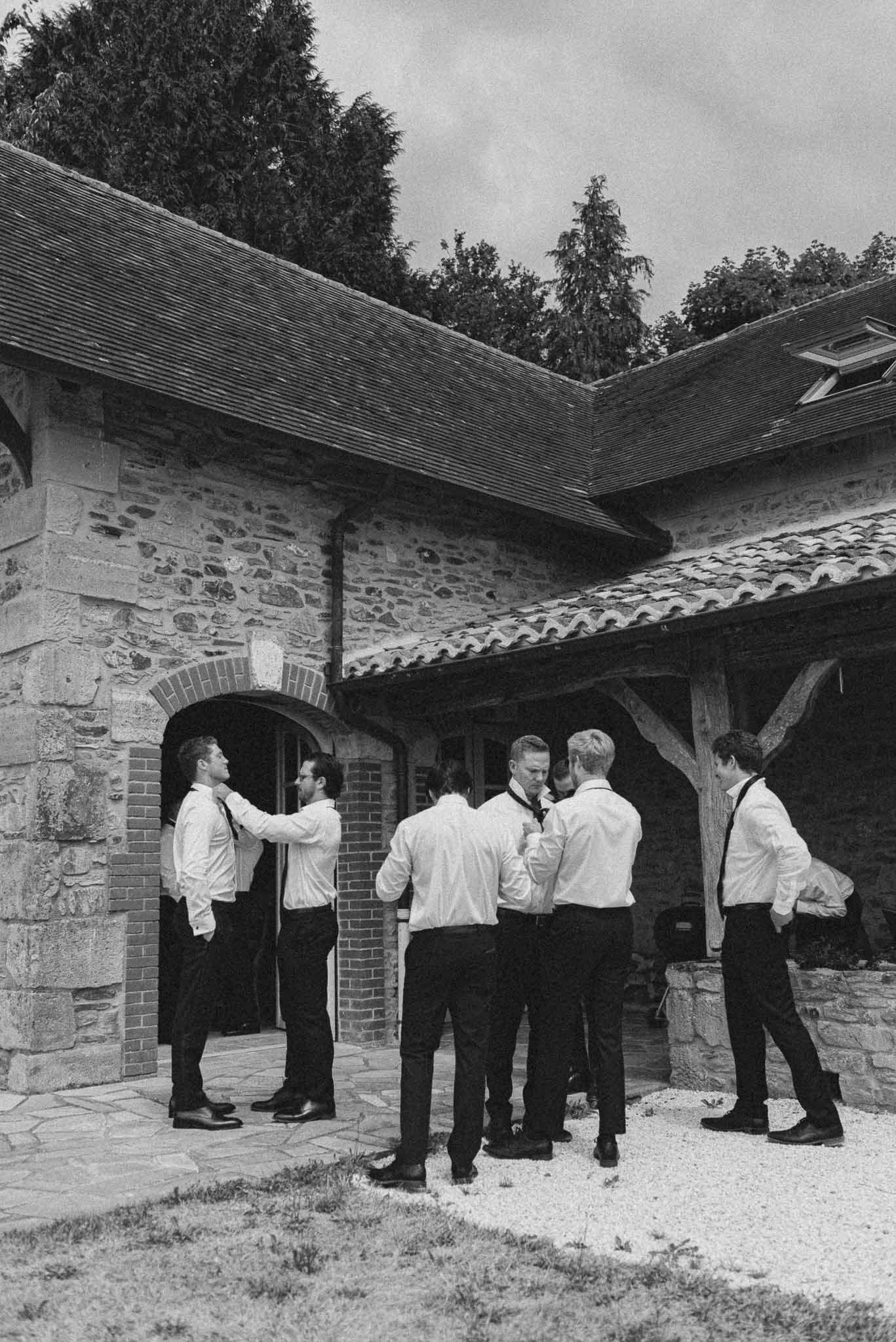 Six groomsmen adjusting ties in white shirts at stone farmhouse courtyard in B&W