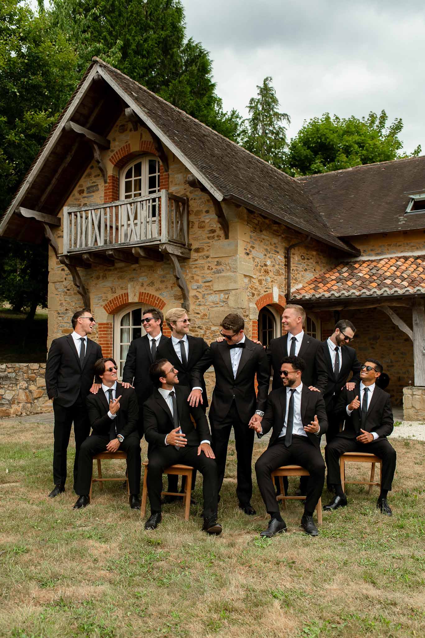 Eleven groomsmen in matching black suits with groom in bow tie seated and standing at stone venue