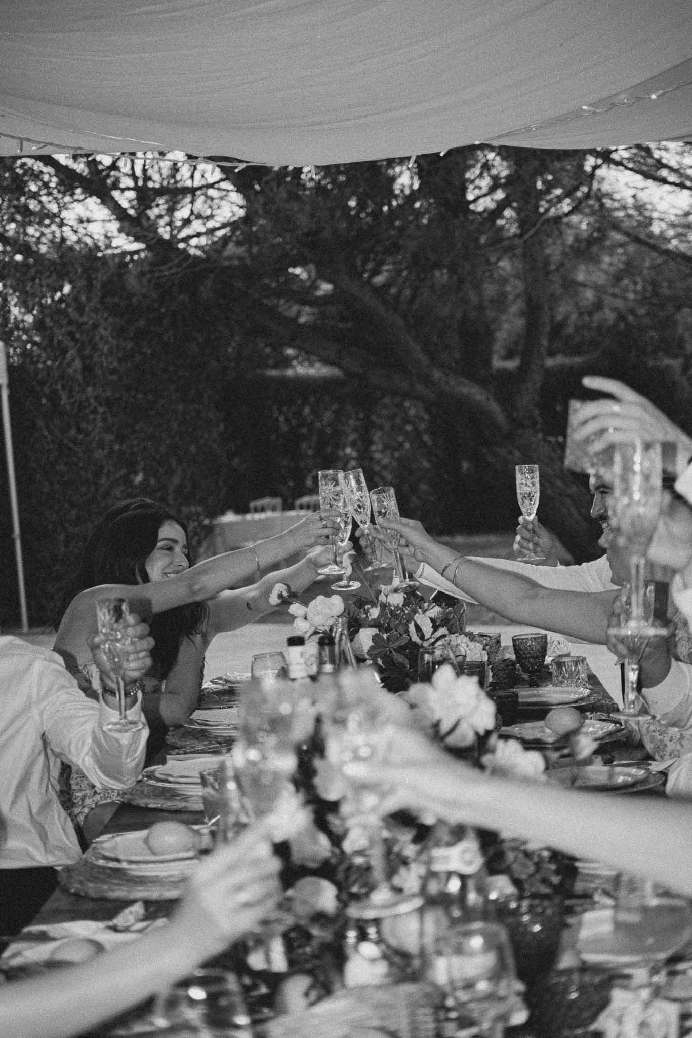 Black-and-white image of guests raising champagne flutes in a toast across a decorated reception table
