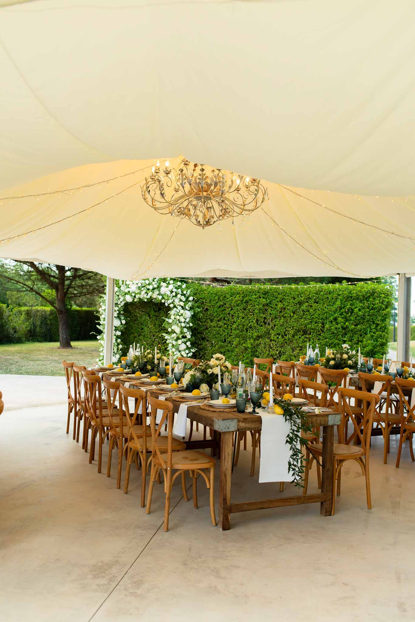 Long wooden farm table under sailcloth tent with greenery garland, lemons, dusty blue glassware, and gold chandelier