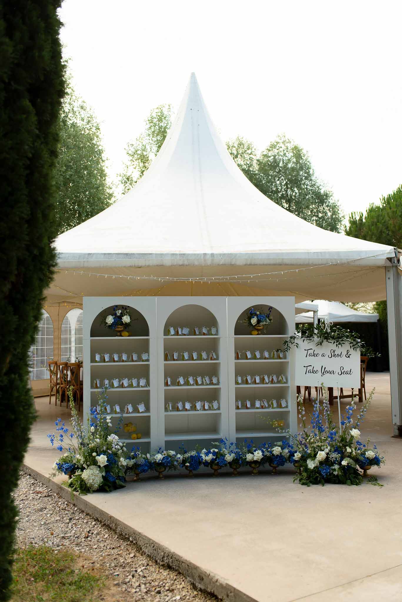 Arched escort card display with shot glass favors, blue hydrangeas and white roses, in front of white marquee tent