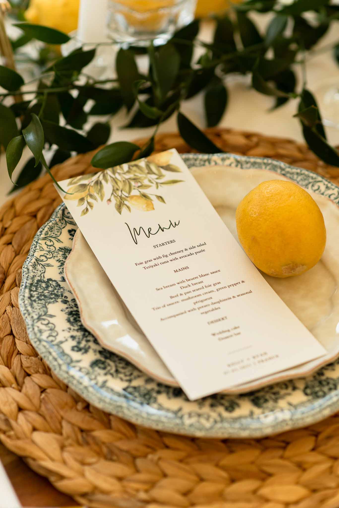 Place setting with rattan charger, toile plate, watercolor lemon menu card, and fresh lemon on white linen