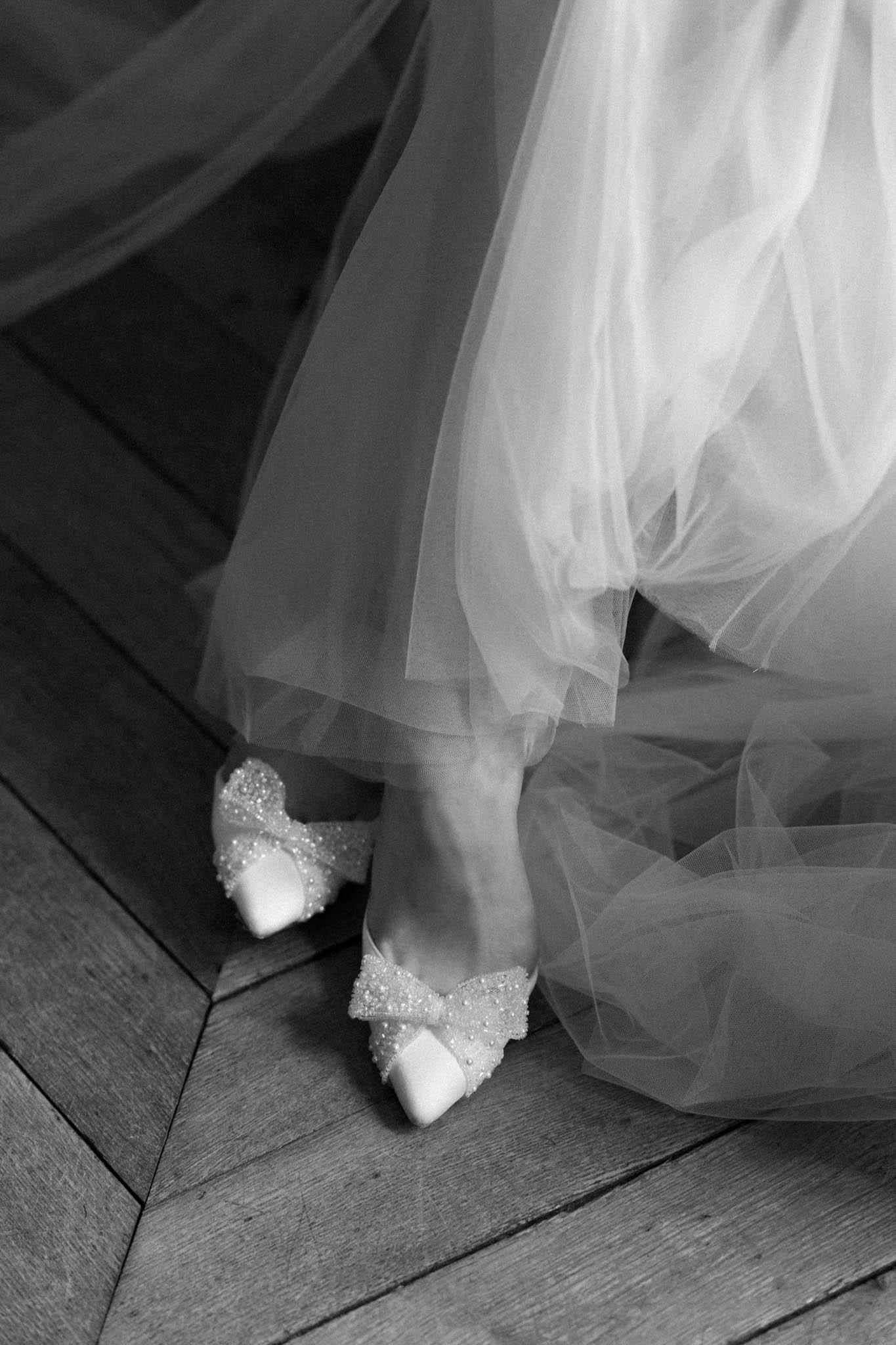 Black and white close-up of crystal bow-embellished pointed heels with pooling tulle on parquet floor