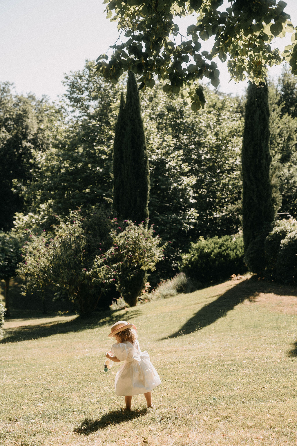 Flower girl in white puff-sleeve dress and pink straw hat running across lawn with cypress trees behind