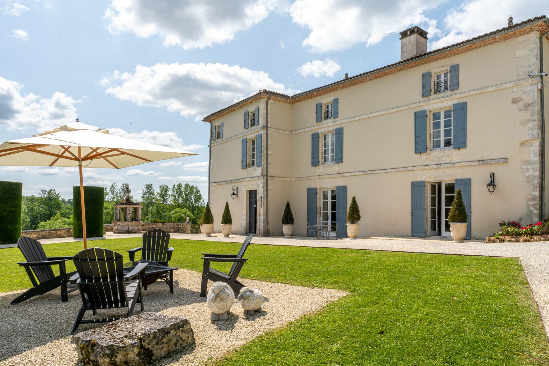 An outdoor wide shot of a French country manor or maison de maître, featuring a cream-rendered three-storey facade with powder blue shutters and French doors. The grounds include a gravel terrace area furnished with black Adirondack chairs grouped around a cream parasol on a wooden frame, two small stone sheep sculptures, and a large natural stone block used as a coffee table. Potted topiary box trees in cream stone urns flank the entrance doors, and a formal hedged garden with a stone fountain is visible in the background to the left. No people are present in the image. Potential venue feature image.