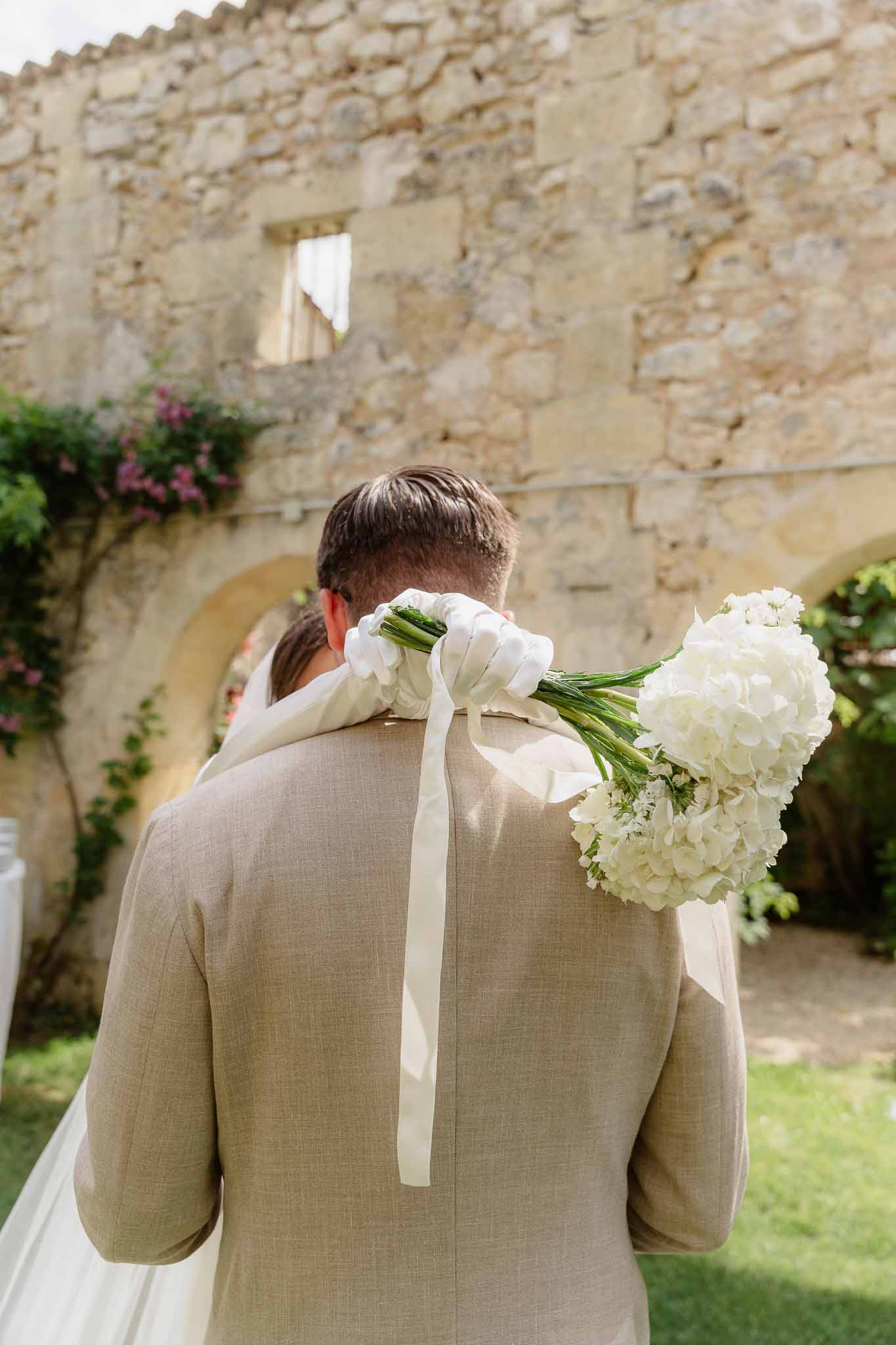 The bride and groom are embracing outdoors in what appears to be a first look or post-ceremony moment, photographed from behind in a close portrait composition. The setting is a stone courtyard or garden area of a French château or mas, with arched stone architecture and climbing plants with pink blooms visible in the background. The groom is wearing a light sand-colored linen suit jacket, and the bride's arm reaches over his shoulder holding a white bouquet of hydrangeas tied with long ivory satin ribbons that trail down the groom's back. The overall styling is minimal and classic, with an all-white and neutral color palette.