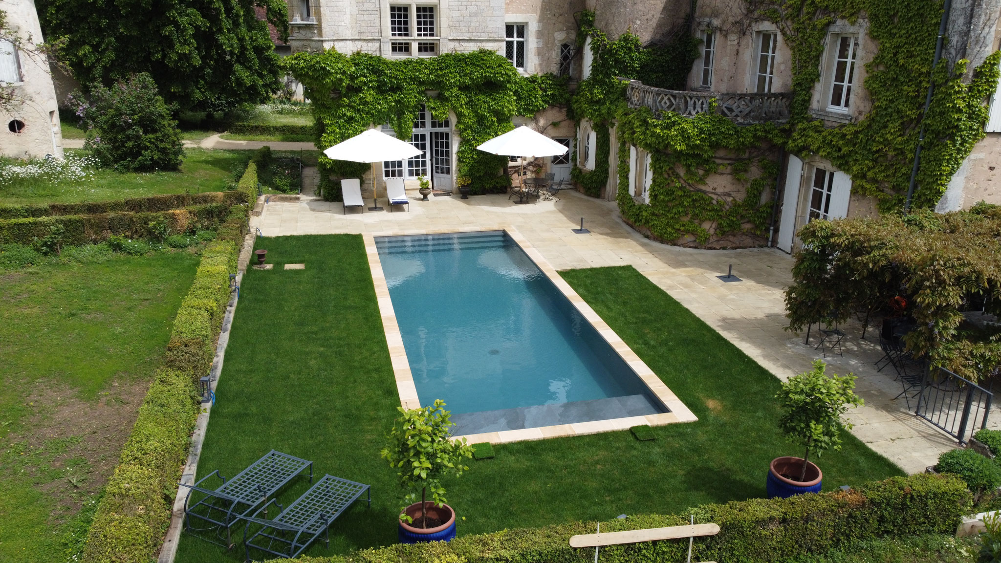 Aerial wide shot of the exterior grounds of a French château venue, showing a rectangular swimming pool with a pale stone surround set within a manicured lawn bordered by trimmed hedges. The château facade is covered in climbing ivy, with tall French windows, a wrought-iron balcony, and white shutters visible. Poolside furniture includes two white market umbrellas, white sun loungers, and dark metal bistro chairs and tables arranged on the stone terrace. Two potted standard trees in cobalt blue ceramic pots are placed near the pool, and additional metal garden benches are positioned at the lower edge of the frame. No people are present. Potential venue feature image.