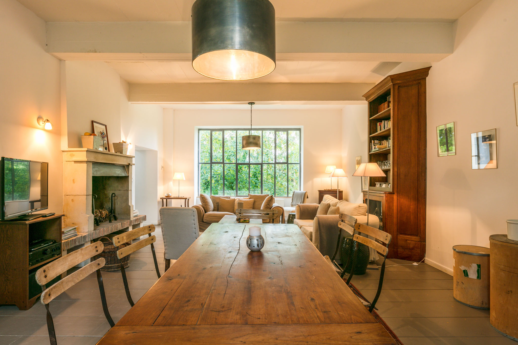 Interior venue shot showing a large open-plan living and dining space inside what appears to be a French country property. The foreground features a long rustic solid-wood farmhouse dining table with mismatched wooden and metal folding chairs, topped with a single metal lantern centerpiece. Overhead hangs a large cylindrical dark metal drum pendant light. The space flows into a sitting area furnished with neutral linen sofas and cushions in sand and taupe tones, arranged around a pallet-style coffee table. A large steel-framed grid window fills the back wall, flanked by table lamps with cream shades. To the left, a stone fireplace mantel with decorative objects sits beside a low wooden media unit with a television. A tall dark walnut armoire bookcase stands to the right alongside framed artwork on white walls. The overall interior palette is warm white, natural wood, and sandy neutrals with a rustic yet refined French country style. Wide-angle interior shot with no people visible. Potential venue feature image.