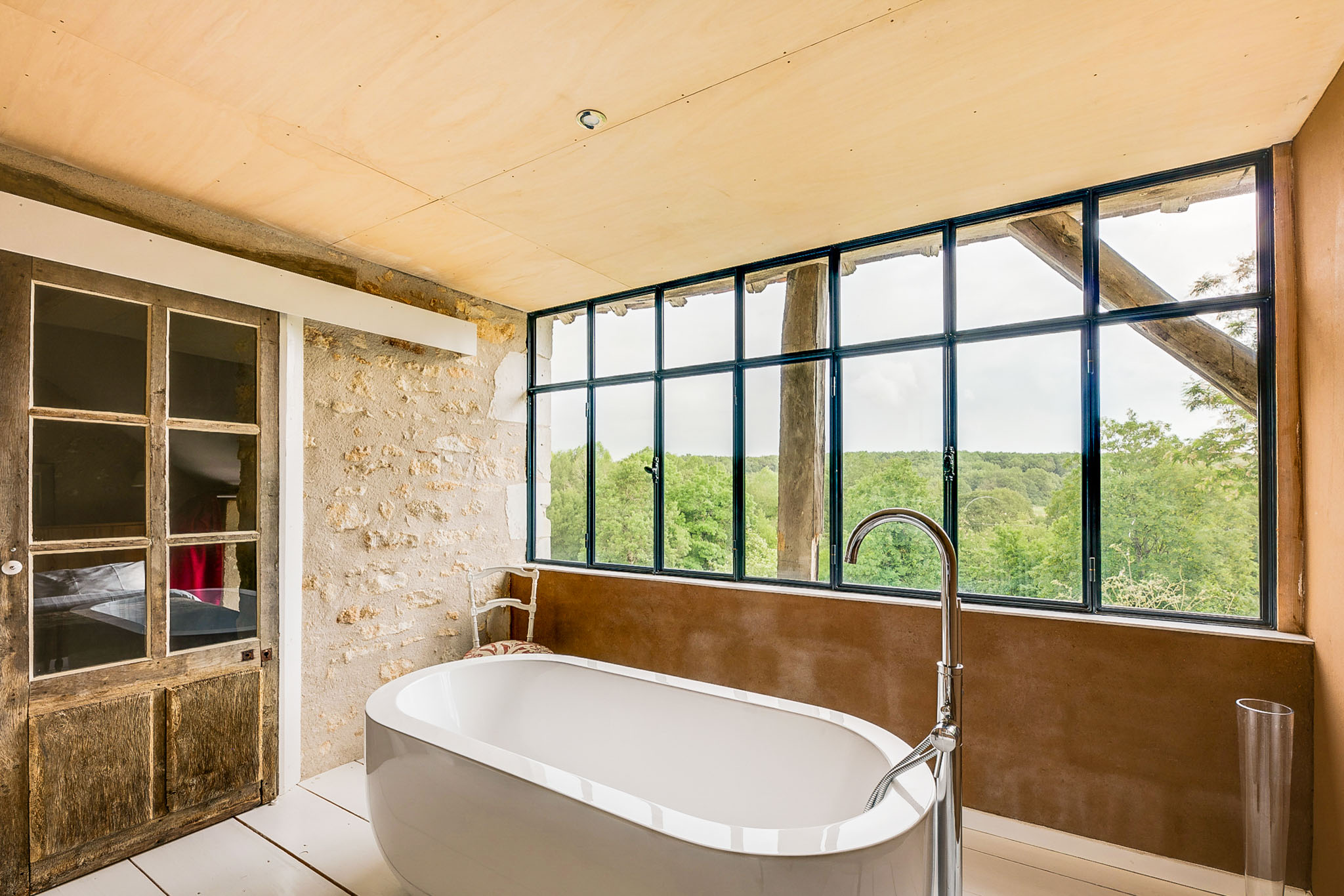Interior wide shot of a bathroom within what appears to be a renovated French rural property. The space features a freestanding white oval soaking tub with a chrome floor-mounted faucet, positioned in front of a large black-framed steel grid window that overlooks a wooded landscape. The design blends old and new: exposed limestone walls and aged wooden double doors with glass panes contrast with the smooth plywood ceiling, white tiled floor, and a rust-toned tadelakt or resin-finished half-wall behind the tub. A vintage upholstered chair is partially visible near the window. The overall aesthetic is rustic-meets-contemporary, with raw heritage materials paired with modern fittings. No people are present. Potential venue feature image.