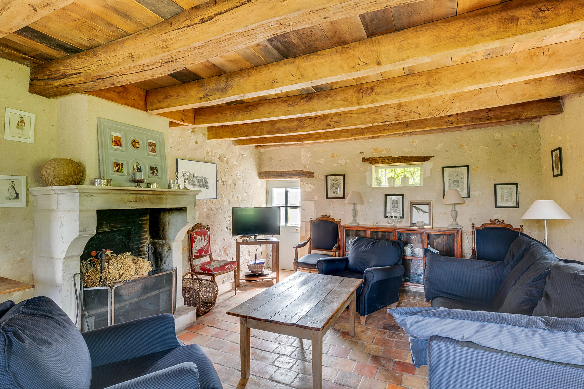 Interior wide shot of a rustic French country living room, likely part of a wedding venue's guest accommodation or common area. The room features exposed heavy wooden ceiling beams, terracotta tile flooring, and bare stone walls with a pale lime render. A large traditional stone fireplace with a decorative mantel holds framed prints and small ornamental objects, with dried flowers and a wicker basket placed in the hearth. Seating includes multiple navy blue upholstered sofas and armchairs arranged around a worn wooden plank coffee table, along with a red-cushioned carved wood accent chair. A dark wood sideboard on the far wall displays framed black-and-white prints, table lamps, and decorative items. The overall style is rustic French farmhouse. Potential venue feature image.
