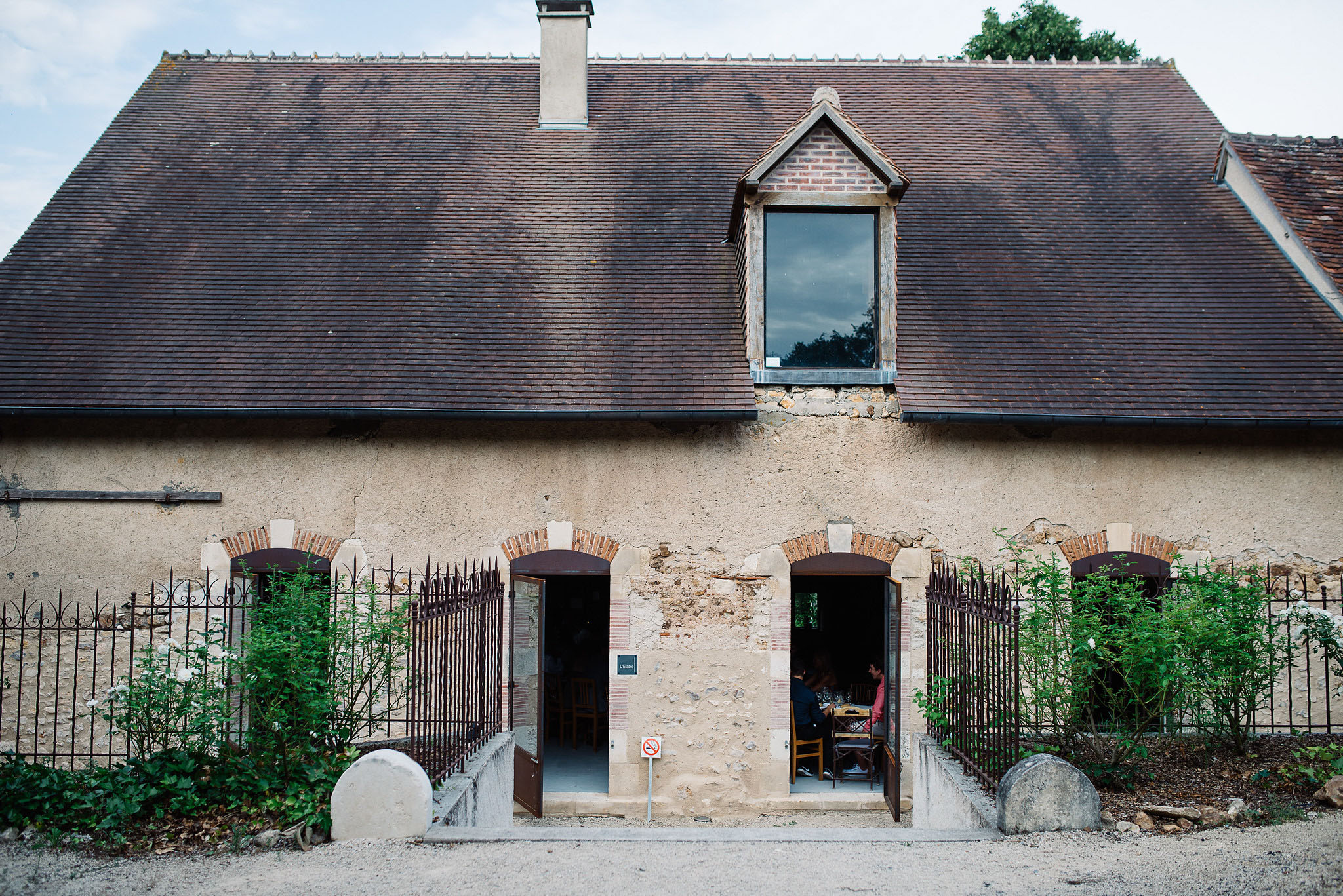 Wide exterior shot of a traditional French rural building featuring a steeply pitched brown tile roof with a central dormer window framed in brick, and a rendered stone facade with arched doorways detailed in red brick and limestone. Two sets of open wooden doors are flanked by wrought iron gates and climbing green shrubs, with rounded stone bollards at the entrance. Through the right-hand doorway, two guests can be seen seated at a wooden table, suggesting a dining or reception space inside. A no-smoking sign is visible near the entrance threshold. Potential venue feature image.