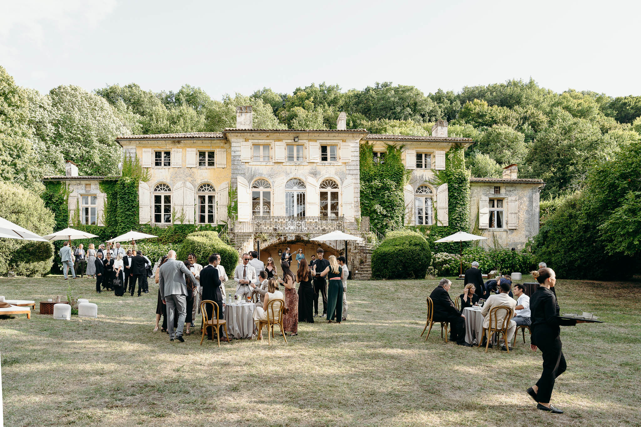 An outdoor cocktail hour is taking place on the lawn in front of a large ivy-covered French manor house with pale limestone facades, arched windows, shutters, and a wrought-iron balcony with exterior stone staircase. Approximately 40–50 guests are mingling across the grounds, seated at round cocktail tables with bentwood chairs or standing in small groups; staff in black uniforms are visible serving guests. White market umbrellas are positioned across the lawn alongside low lounge seating with white cushions on the left side. Guests are dressed in formal attire — suits in grey, navy, and black, alongside floor-length gowns in black, dark green, and floral prints — suggesting a classic, formal wedding aesthetic. The wide-shot composition captures both the full architecture of the venue and the full spread of the cocktail hour activity. Potential venue feature image.