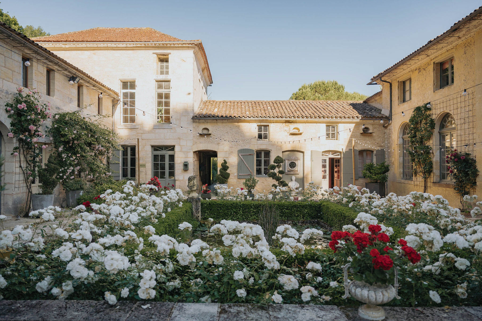 Wide shot of a Provençal-style stone courtyard venue featuring honey-colored limestone buildings with terracotta-tiled roofs, grey-painted shutters, and arched windows. The foreground is filled with densely planted white and red roses in formal garden beds bordered by low boxwood hedging, with a stone urn planter and classical bust sculptures visible among the plantings. Strings of bistro lights are strung across the courtyard between the buildings, and climbing roses in pink and red grow against the façades. No people are present in the image. Potential venue feature image.