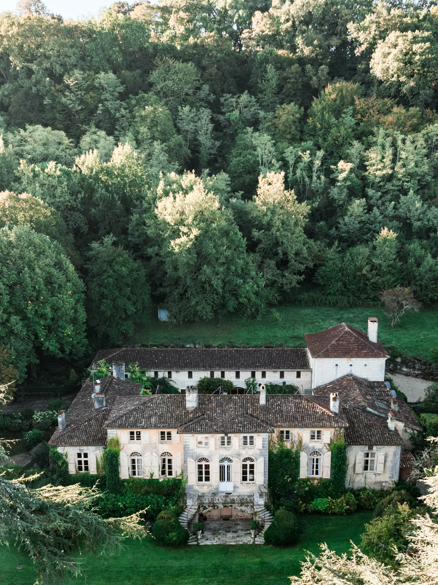 An aerial drone shot of a French country estate, showing a large multi-wing stone manor house with terracotta-tiled roofs, arched fanlight windows, wooden shutters, and a grand front staircase with wrought iron railings. The main building is flanked by outbuildings and a secondary structure with a red-tiled hip roof and chimney, all in pale limestone. Climbing ivy and clipped topiary cover portions of the facade, and a manicured lawn extends in front of the property. The entire estate is set against a dense backdrop of mature woodland. No people are visible in the image. Potential venue feature image.