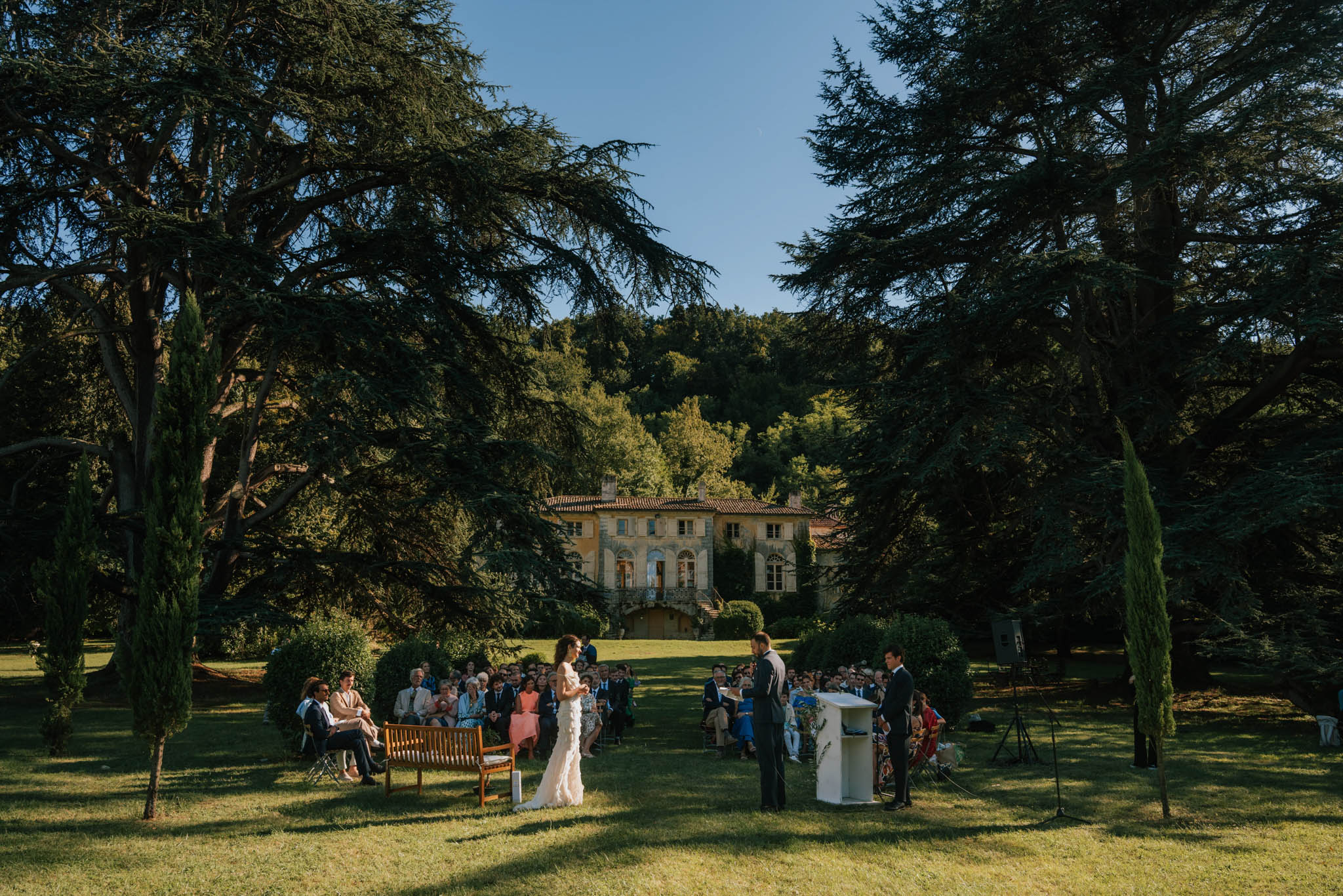 An outdoor wedding ceremony taking place on a wide lawn in front of a large French manor house or château with a terracotta roof, arched windows, and a balcony with exterior staircases. The bride, wearing a white fitted gown with ruffle or lace detailing, stands facing the groom, who is dressed in a dark suit, with an officiant at a white pedestal lectern to the right. Approximately 50–70 seated guests are arranged in rows on the lawn, with a wooden bench visible at the front left. The wide-angle shot frames the ceremony between large cedar trees and tall cypress trees, with the manor facade visible in the background mid-distance. The ceremony has a classic, unfussy outdoor style with no visible floral arches or decor beyond the white lectern. Potential venue feature image.