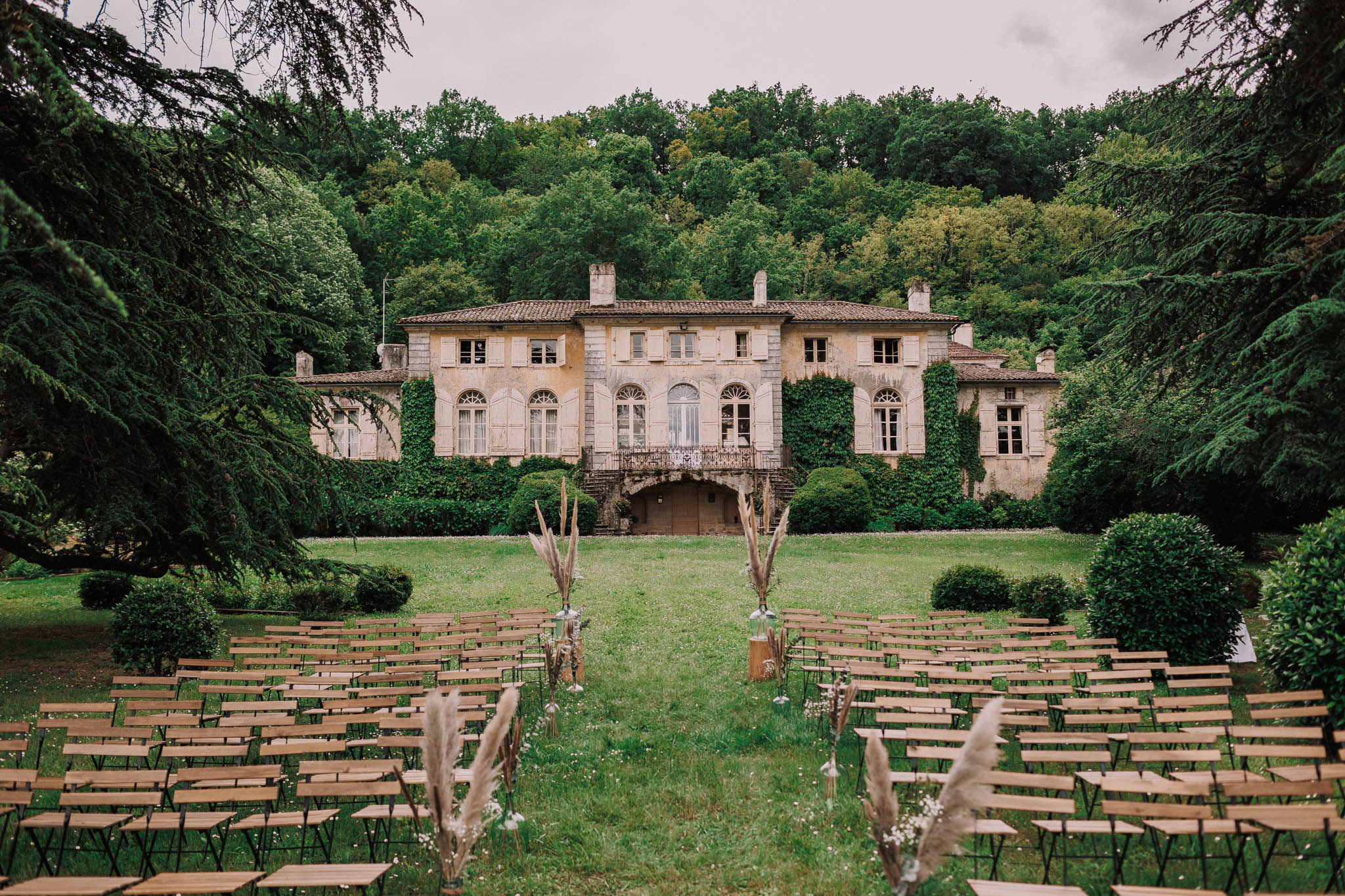 An outdoor wedding ceremony setup on the lawn of a large French manor house or château, with no guests or couple present — the space is arranged and awaiting the ceremony. Rows of light wood folding chairs with black metal frames are arranged in two sections separated by a central grass aisle. The aisle is lined with dried pampas grass and small glass bud vases, along with taller arrangements of dried grasses and delicate white wildflowers placed on wooden pedestals at intervals. The décor palette is natural and earthy, leaning toward a boho-rustic style with dried botanical elements rather than fresh floral arrangements. The manor building in the background is a two-storey stone structure with ivy-covered facades, arched windows with shutters, a wrought-iron balcony, and a tiled roof. Wide shot taken from behind the guest seating looking toward the house. Potential venue feature image.