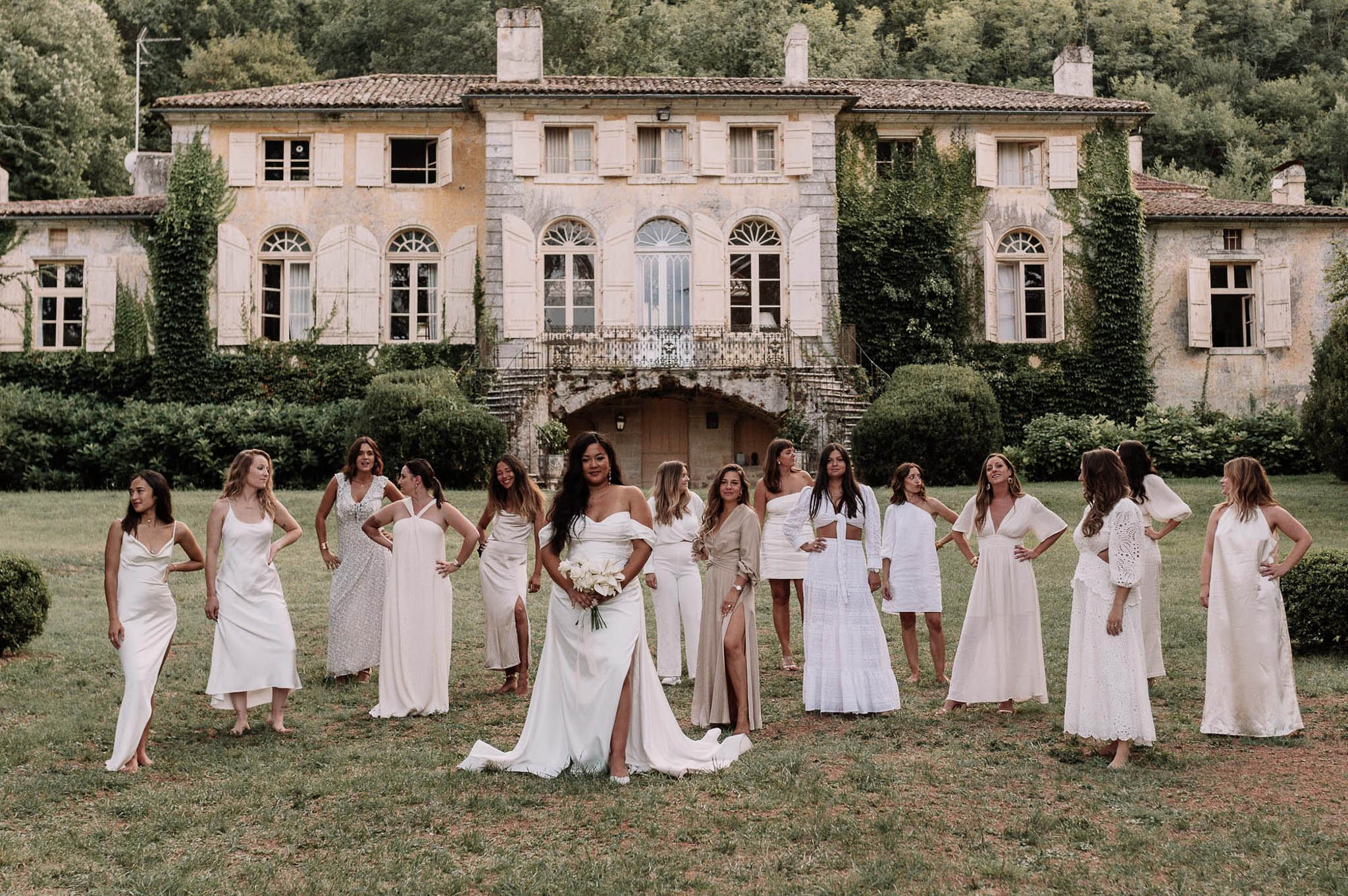 A bridal party group portrait taken outdoors on the lawn of a large French manor or bastide, featuring the bride and approximately twelve bridesmaids arranged in a loose arc. The bride stands at center, wearing an off-the-shoulder white gown with a front slit and train, holding a bouquet of white calla lilies or tulips. The bridesmaids wear a mix-and-match palette of white, ivory, and champagne-beige outfits in varied styles including satin slip dresses, lace midi dresses, a two-piece crop top set, a sheer coverup, and a structured mini dress, creating a cohesive but individualized look. The venue is a multi-story Provençal-style manor with arched windows, white shutters, ivy-covered facades, a wrought-iron balcony, and a grand stone entrance staircase. The overall styling theme is modern boho with a white and neutral color palette. Wide shot composition.