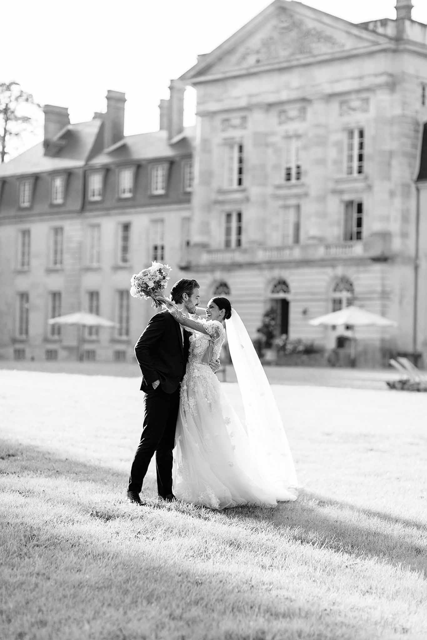 This is a black-and-white couple portrait taken outdoors on the grounds of a large French château. The bride and groom are embracing and laughing together on a lawn in front of the main façade of the château, which features classical French architecture with a pediment, tall windows, and stone masonry rendered in bright mid-tones against a high-key sky. The groom wears a dark suit, while the bride wears a long-sleeved, heavily lace-appliquéd ball gown with a full tulle skirt and a cathedral-length veil that trails behind her in the light breeze. She holds a full, rounded bouquet. The image is a full-length portrait shot with the château softly out of focus in the background, creating strong depth of field with high contrast between the dark foreground lawn and the bright building facade. Potential venue feature image.