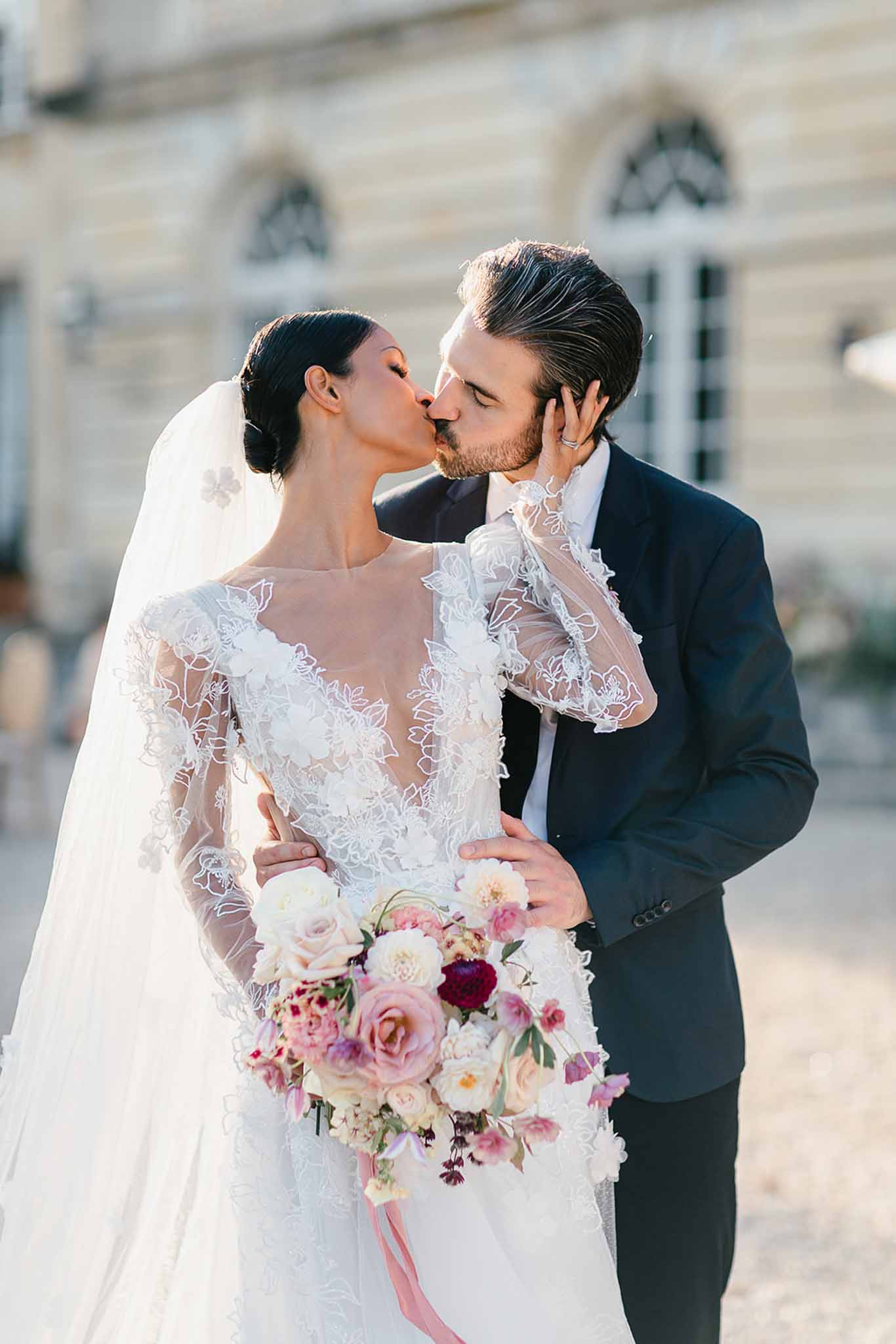 A couple portrait shot outdoors in front of a classical French stone building with arched windows, captured in warm natural light. The bride and groom are kissing, with the bride's hand resting on the groom's face. The bride wears a long-sleeve illusion lace gown with floral appliqué detailing over a sheer bodice, her dark hair styled in a low bun with a cathedral-length veil featuring floral appliqués. The groom wears a fitted navy suit with a white shirt. The bride holds a large cascading bouquet composed of blush roses, cream peonies, deep burgundy dahlias, mauve and dusty pink garden roses, and trailing pink ribbon. The overall styling is classic and modern, with a romantic color palette of blush, burgundy, and cream. Medium portrait shot with a shallow depth of field, background softly blurred.