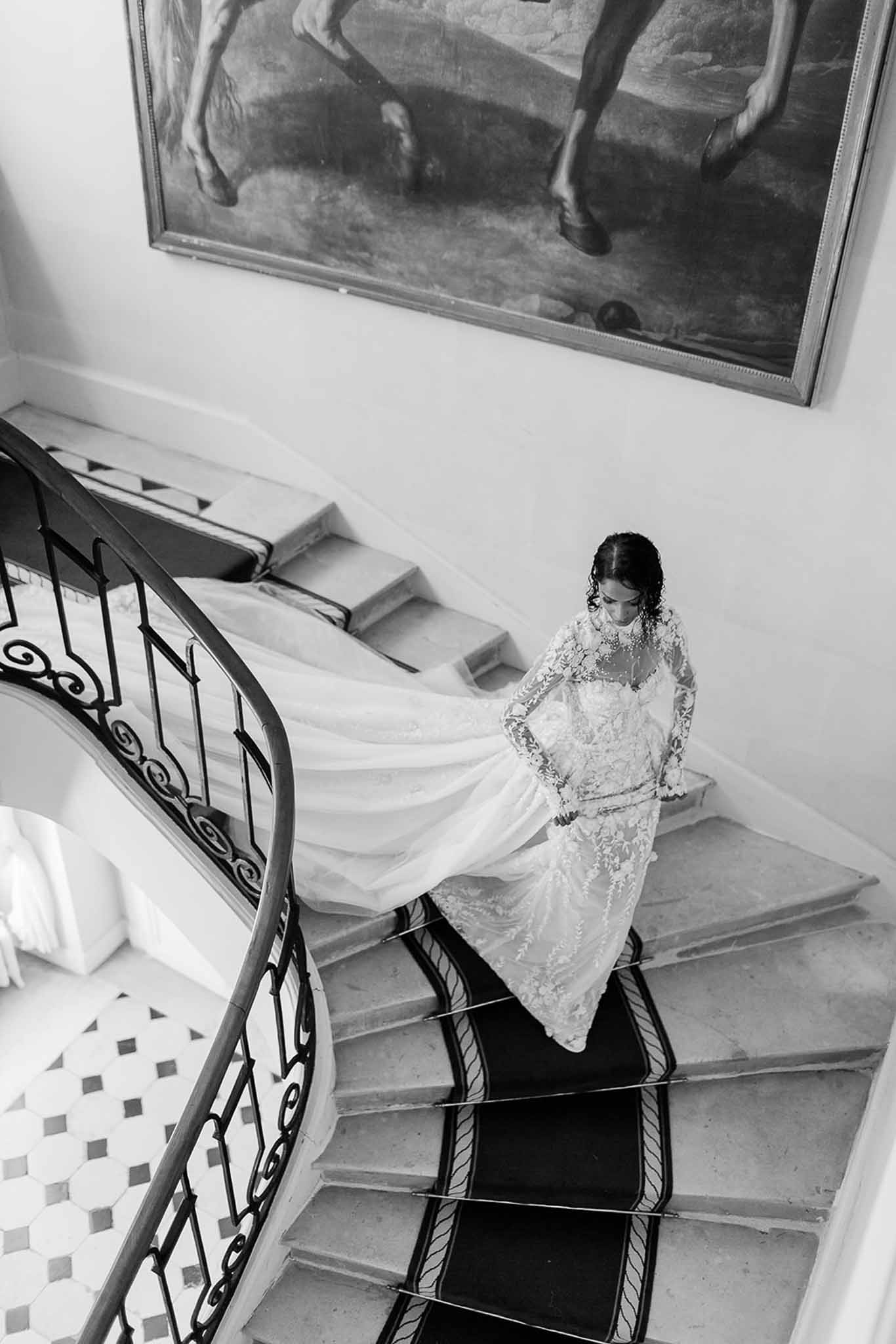 This black-and-white bridal portrait shows a bride descending a grand curved marble staircase inside what appears to be a chateau or manor house. The image is shot from above, creating a dramatic overhead composition that emphasizes the full sweep of the bride's cathedral-length train spreading across the steps. She wears a long-sleeve, high-neck lace wedding gown with intricate floral lace detailing on the bodice and sleeves, paired with a voluminous tulle skirt. The staircase features wrought-iron scrollwork balustrades, a dark carpet runner along the center, and a black-and-white geometric tile floor visible at the base; a large framed oil painting depicting a horse hangs on the wall above the landing. The high-contrast black-and-white tones give strong definition to the lace texture and the sweeping architectural lines of the interior.