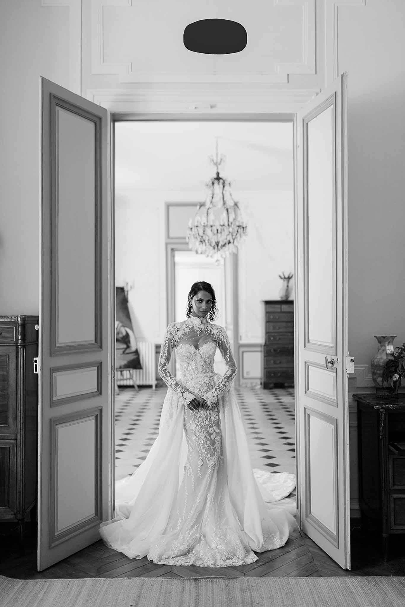 Black-and-white bridal portrait taken indoors at what appears to be a French château or manor house. The bride stands centered in a set of tall double doors with panelled woodwork, facing the camera with her hands clasped at her waist. She wears a fitted long-sleeve lace gown with a high illusion neckline, intricate floral appliqué detail throughout the bodice and sleeves, and a flowing train that extends onto the herringbone parquet floor in front of her. The room behind her features a diamond-pattern black-and-white tiled floor, a crystal chandelier, a tall gilt-framed mirror, and antique dark wood furniture, conveying a classic French interior aesthetic. The image is a full-length portrait shot with strong contrast between the bright interior beyond the doorway and the darker foreground framing.