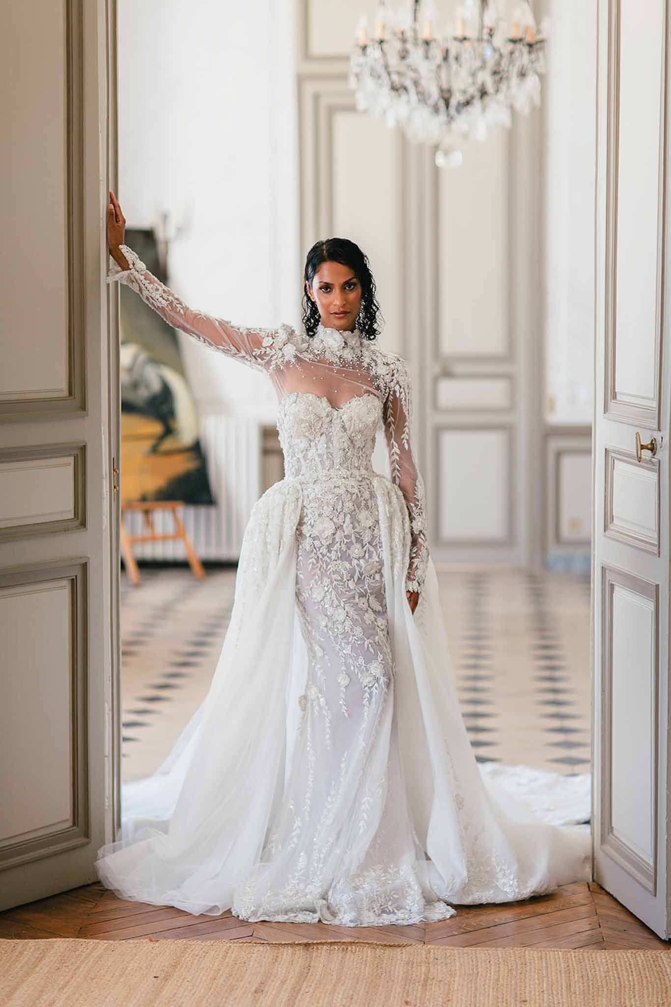 A bridal portrait of a single bride standing in a set of open double doors inside what appears to be a French château or manor house. The bride wears a white fitted lace gown with sheer long sleeves, a high neckline adorned with 3D floral appliqués, and an attached overskirt or detachable tulle layer that creates volume at the hips and flows into a full train. The bodice features heavily embroidered floral and leaf motifs with what appear to be pearl or crystal embellishments. Her dark hair is pulled back, and she leans one arm against the door frame while facing the camera directly. The interior setting features panelled grey-painted doors, a crystal chandelier, herringbone parquet flooring, and a geometric tile floor visible through the doorway — consistent with a classic French period interior. The styling is formal and high-fashion. Full-length portrait shot with the doorframe used as a natural frame within the composition.