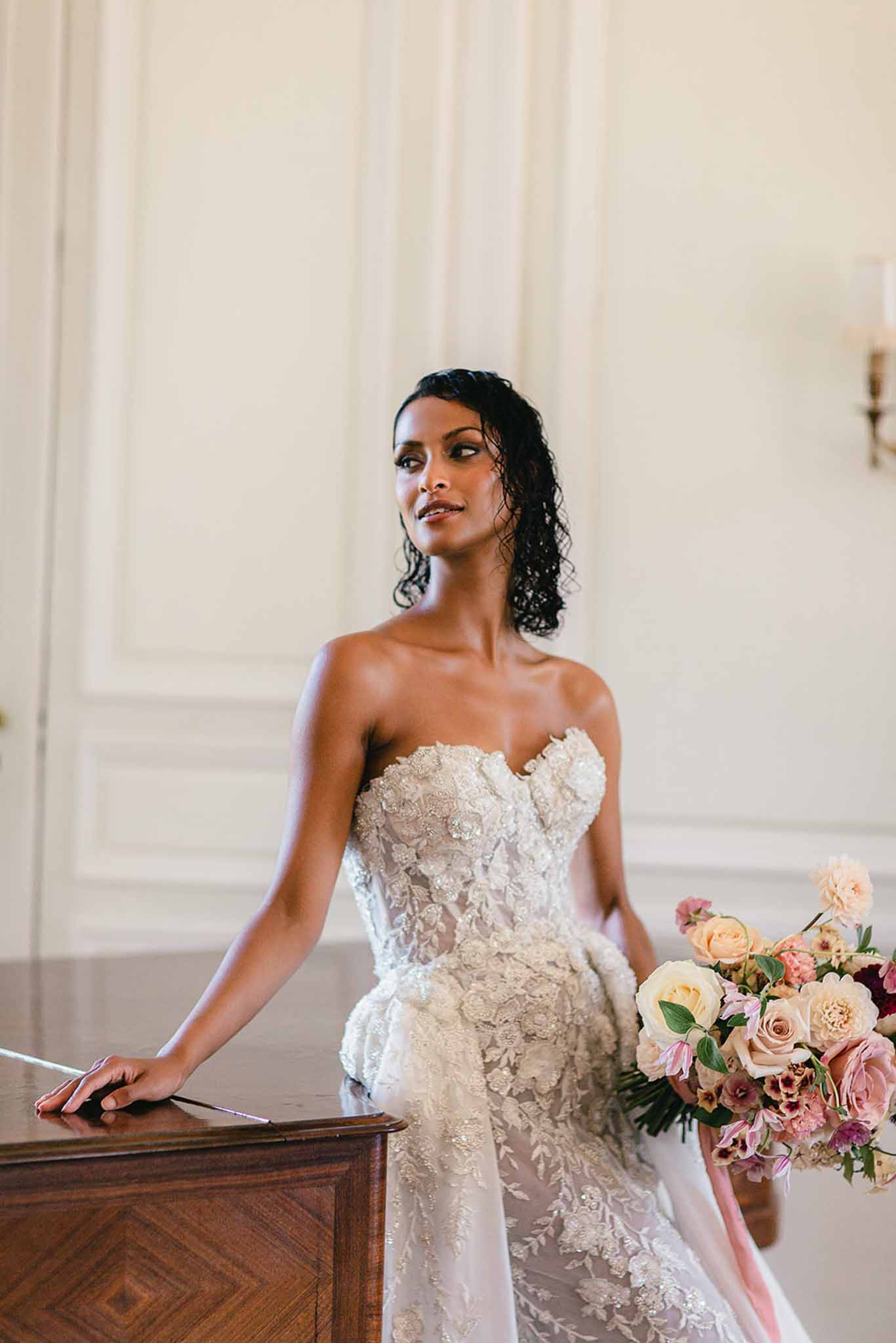A bridal portrait shot indoors, set against cream-painted wood-panelled walls with a wall sconce visible in the background, suggesting a classic French château or manor interior. The bride is seated and leaning against a dark wood antique desk or console, gazing to one side, with her dark curly hair styled half-up. She wears a strapless sweetheart-neckline gown heavily embellished with beaded and embroidered floral lace appliqués in ivory and champagne tones. She holds a large, loosely arranged bouquet featuring blush roses, peach roses, mauve and dusty rose blooms, burgundy accents, and trailing blush ribbon, with green foliage throughout. The composition is a mid-length portrait with a soft, airy feel, and the styling leans toward classic with a romantic, ornate edge.