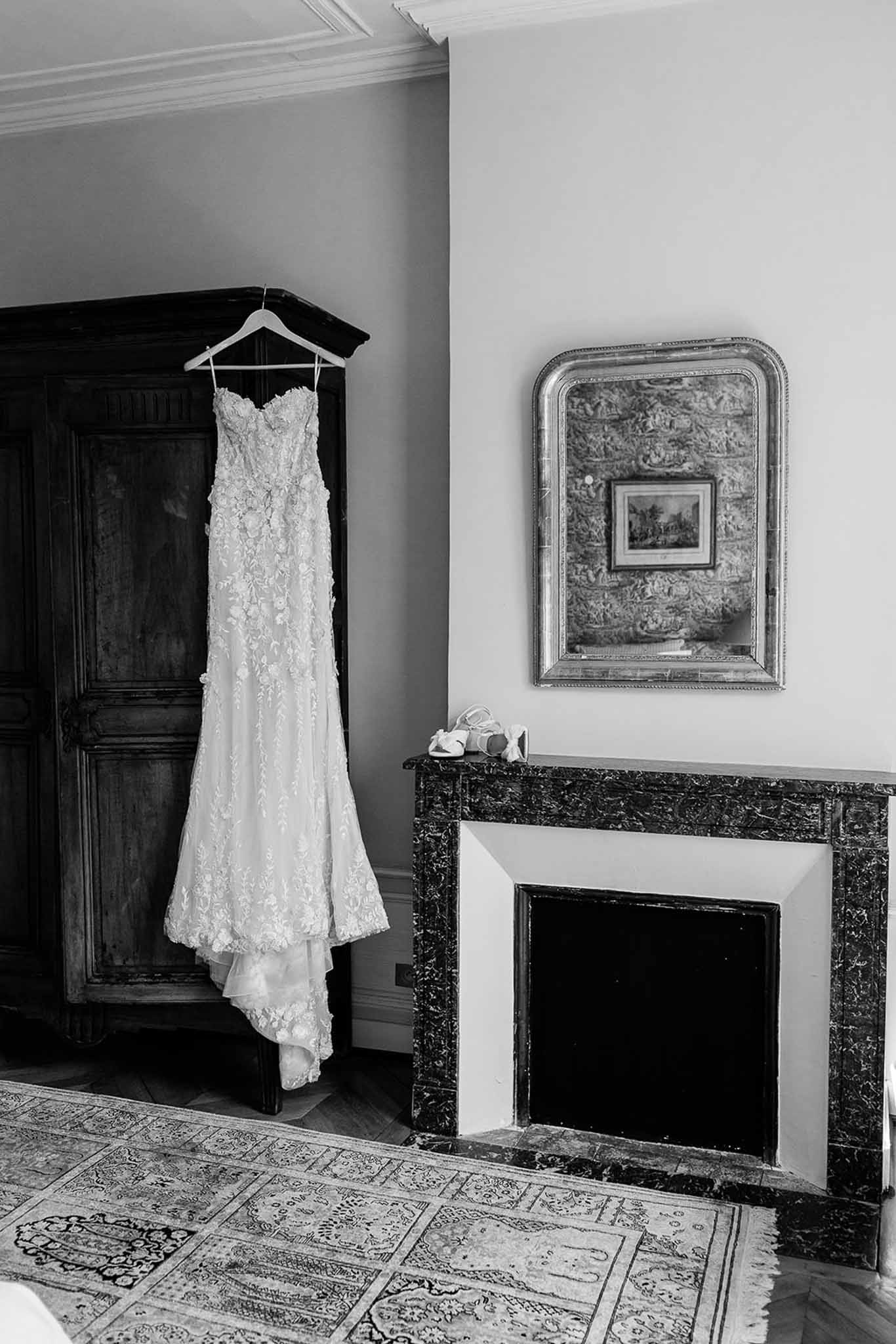 This is a black-and-white getting-ready detail shot taken indoors in what appears to be a classic French château bedroom or salon. A strapless, floor-length bridal gown with dense floral lace appliqué and a fitted silhouette hangs from a hanger on the cornice of a large, dark antique wooden armoire. To the right, a dark marble fireplace mantel holds a pair of strappy bridal heels, and an ornate gilt-framed mirror hangs on the wall above it, reflecting a framed artwork. A patterned area rug covers the parquet floor in the foreground. The image uses strong contrast between the dark wood furniture and the light-toned dress and walls, giving the scene a classic, formal feel.