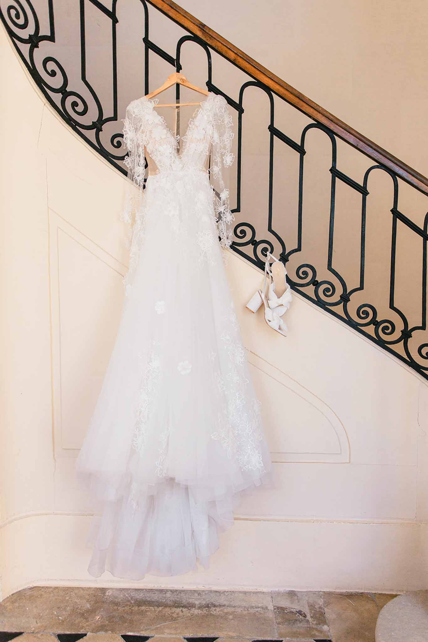 A getting-ready detail shot of a white bridal gown hanging from a wooden hanger on a curved interior staircase railing with black wrought-iron scrollwork. The gown features a V-neckline, long sheer sleeves with 3D floral appliqué detailing, lace embroidery across the bodice and skirt, and a layered tulle A-line silhouette. A pair of white block-heel ankle-strap sandals is hooked onto the railing beside the dress. The setting is an indoor stairwell with cream-painted panelled walls and a warm wood handrail, consistent with a classic French château or manor house interior. The composition is a full-length vertical portrait shot of the dress styled against the architectural backdrop.