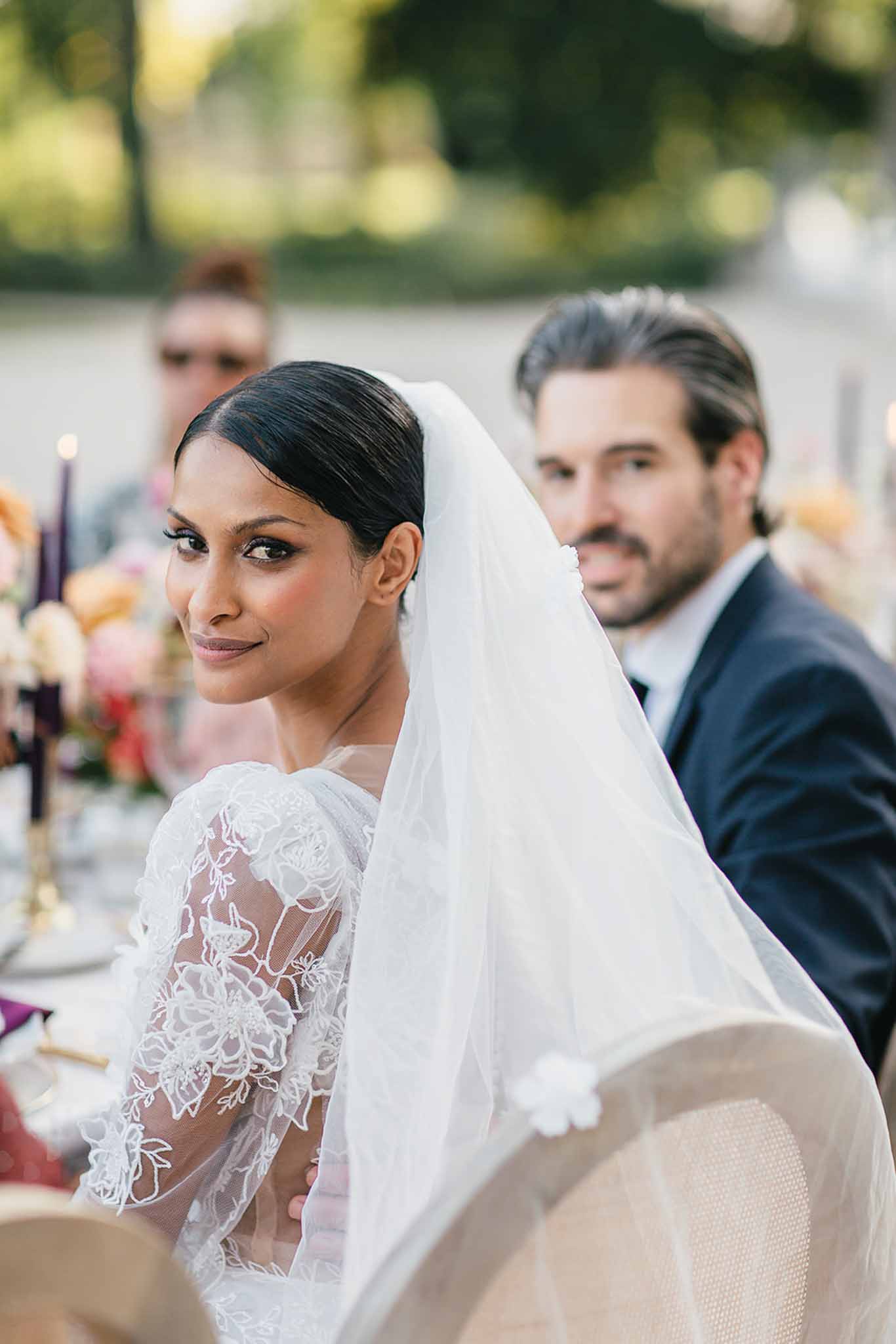 The bride is seated at the wedding reception dinner table outdoors, turning to look over her shoulder toward the camera, with the groom visible and slightly out of focus behind her in a navy suit. She is wearing a long-sleeve white lace gown with floral embroidery detailing and a flowing veil trimmed with a small white flower accent, with her dark hair pulled back sleekly. The table setting in the background features tall purple taper candles in gold holders, and colorful floral arrangements in peach, coral, and pink tones, suggesting a rich, jewel-toned decor palette. The shot is a close-up portrait taken at an outdoor reception, with soft warm natural light and a shallow depth of field blurring the background guests and greenery.