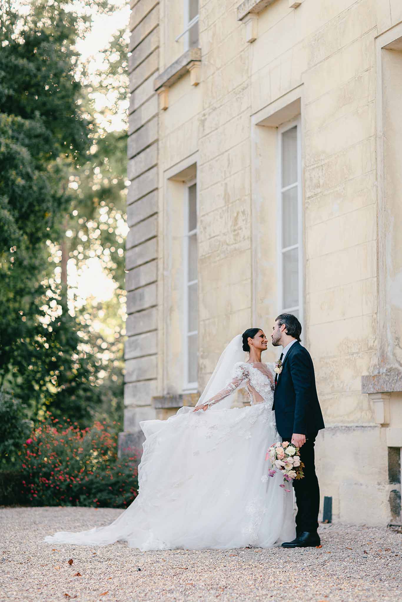 A couple portrait taken outdoors against the limestone facade of a French château, shot at golden hour. The bride wears a full-volume white ballgown with long lace-embroidered illusion sleeves and a long cathedral veil, and holds a bouquet of blush and ivory blooms with greenery; the groom wears a navy suit with a white boutonnière. The two are standing close together, faces nearly touching, on a gravel path alongside the château exterior. The composition is a full-length wide portrait with warm, soft backlighting filtering through the trees to the left.
