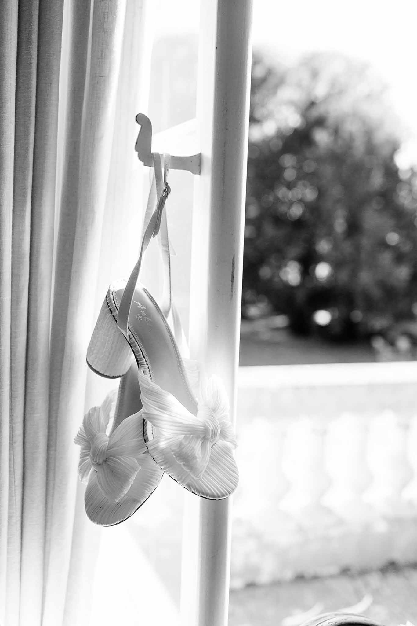 A black-and-white close-up detail shot of a pair of bridal heeled sandals hanging from a white hook on a door or window frame during the getting-ready phase. The shoes feature a textured, pleated fabric construction with a knotted toe strap detail and a block heel, and appear to be white or ivory in tone. Sheer curtains frame the left side of the image, and a softly blurred outdoor scene is visible through a window in the background, creating bright, diffused natural backlighting. The composition is a portrait-orientation detail shot with strong contrast between the light shoes and the bright window light.