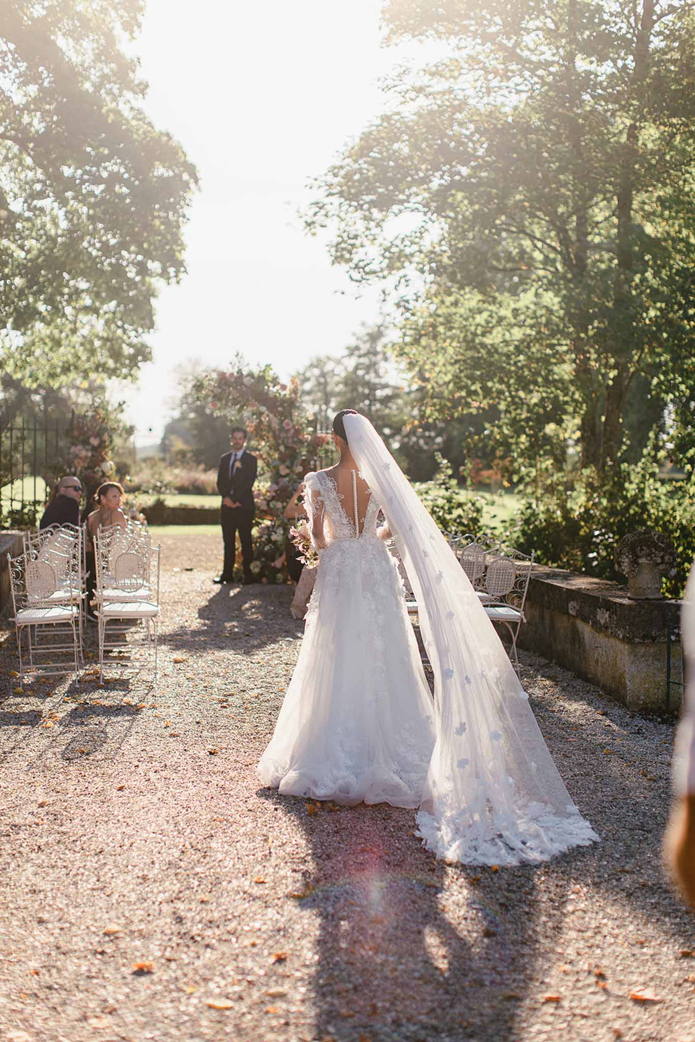 The bride walks down the aisle during an outdoor garden ceremony, photographed from behind as she approaches the groom who stands waiting at the altar. She wears a full A-line white gown with a deep V-back, long lace-appliquéd sleeves, and a voluminous skirt adorned with 3D floral appliqués; her cathedral-length veil is embellished with scattered floral or butterfly appliqués and fans out across the gravel path. The ceremony is set in a formal garden with a gravel aisle, ornate white wrought-iron chairs seating a small group of guests on the left, and a floral arch decorated with blush, peach, and terracotta toned blooms framing the altar in the background. The image is a wide shot taken at ground level with strong backlighting creating a warm lens flare, giving the scene a bright, hazy quality.