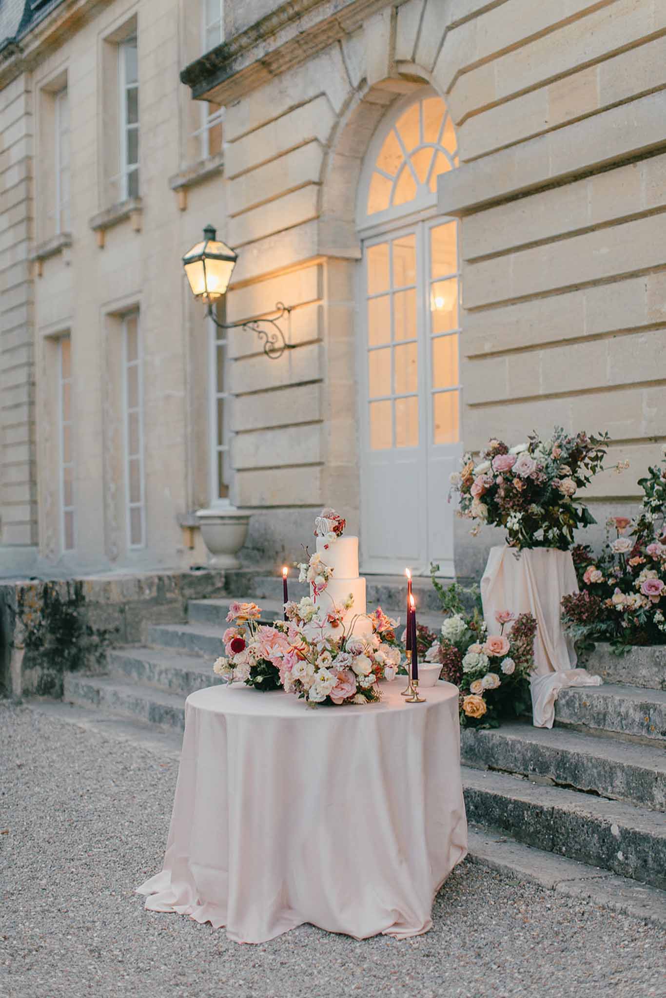 A wedding cake display set outdoors at dusk in front of a classic French château facade with tall arched windows and warm interior lighting glowing through the glass. A four-tier smooth ivory cake sits on a round table draped in a blush pink satin linen, surrounded by a lush arrangement of blush pink, coral, cream, and burgundy flowers including roses, ranunculus, and garden blooms. Two deep burgundy taper candles in gold candlestick holders flank the cake, both lit. To the right, a large floral installation in matching blush, coral, cream, and burgundy tones is displayed on a pedestal draped in ivory fabric at the base of stone steps. A wall-mounted iron lantern above adds ambient warm light to the scene. The overall styling palette combines blush, burgundy, and ivory in a classic, romantic outdoor setting. Wide shot composition. Potential venue feature image.