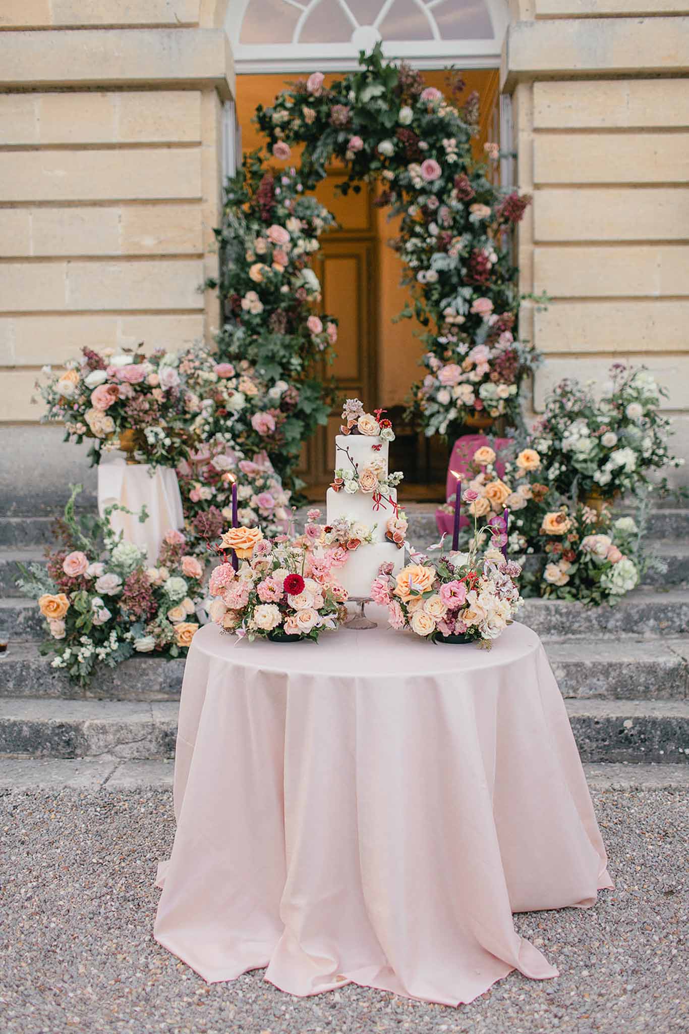 A cake display setup positioned outdoors on the stone steps and forecourt of a classical French château or manor, with pale limestone architecture visible in the background. A three-tiered white wedding cake sits on a round table draped in a blush pink linen, flanked by two low floral arrangements and two lit deep purple taper candles; the cake itself is decorated with applied sugar or fresh flowers including peach roses, pink blooms, and small red accents. Surrounding the entrance doorway behind the table is a large circular floral arch constructed from lush greenery, blush pink roses, peach garden roses, dusty mauve blooms, and burgundy accents, with additional freestanding floral column arrangements on either side of the steps in the same palette. The overall color palette combines blush, peach, dusty rose, burgundy, and deep plum against green foliage, creating a romantic garden-inspired aesthetic. Wide shot, centered composition. Potential venue feature image.